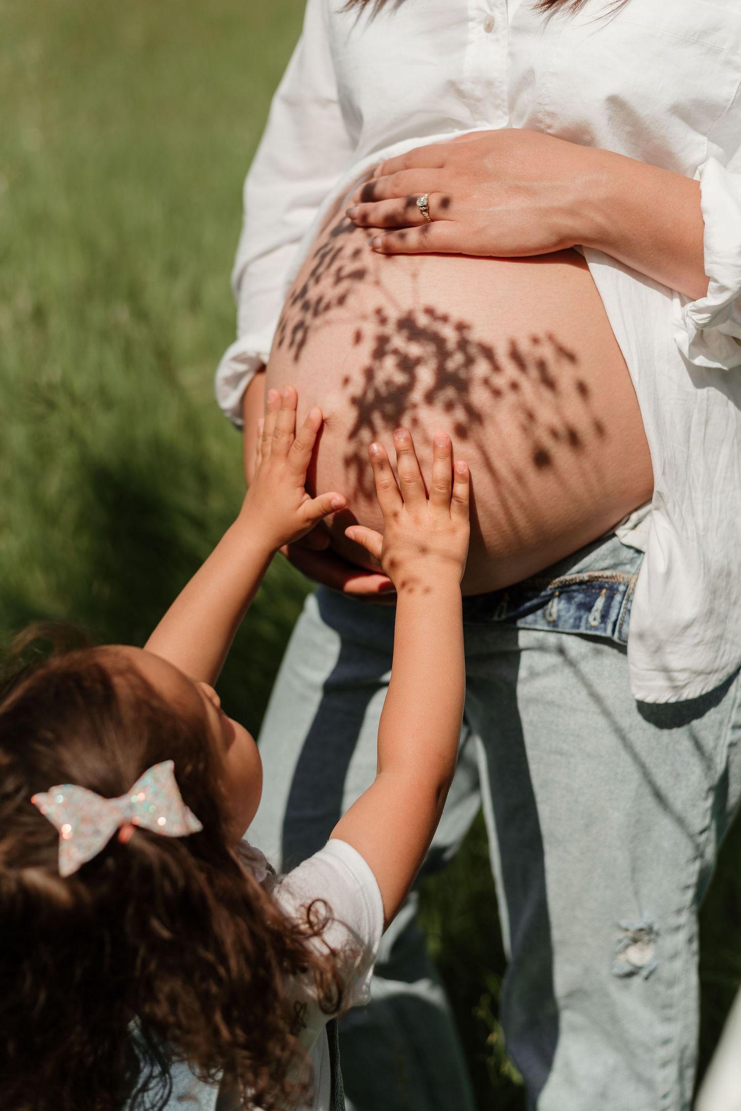 Small hands reach up to touch a pregnant belly with dappled sunlight creating shadows.