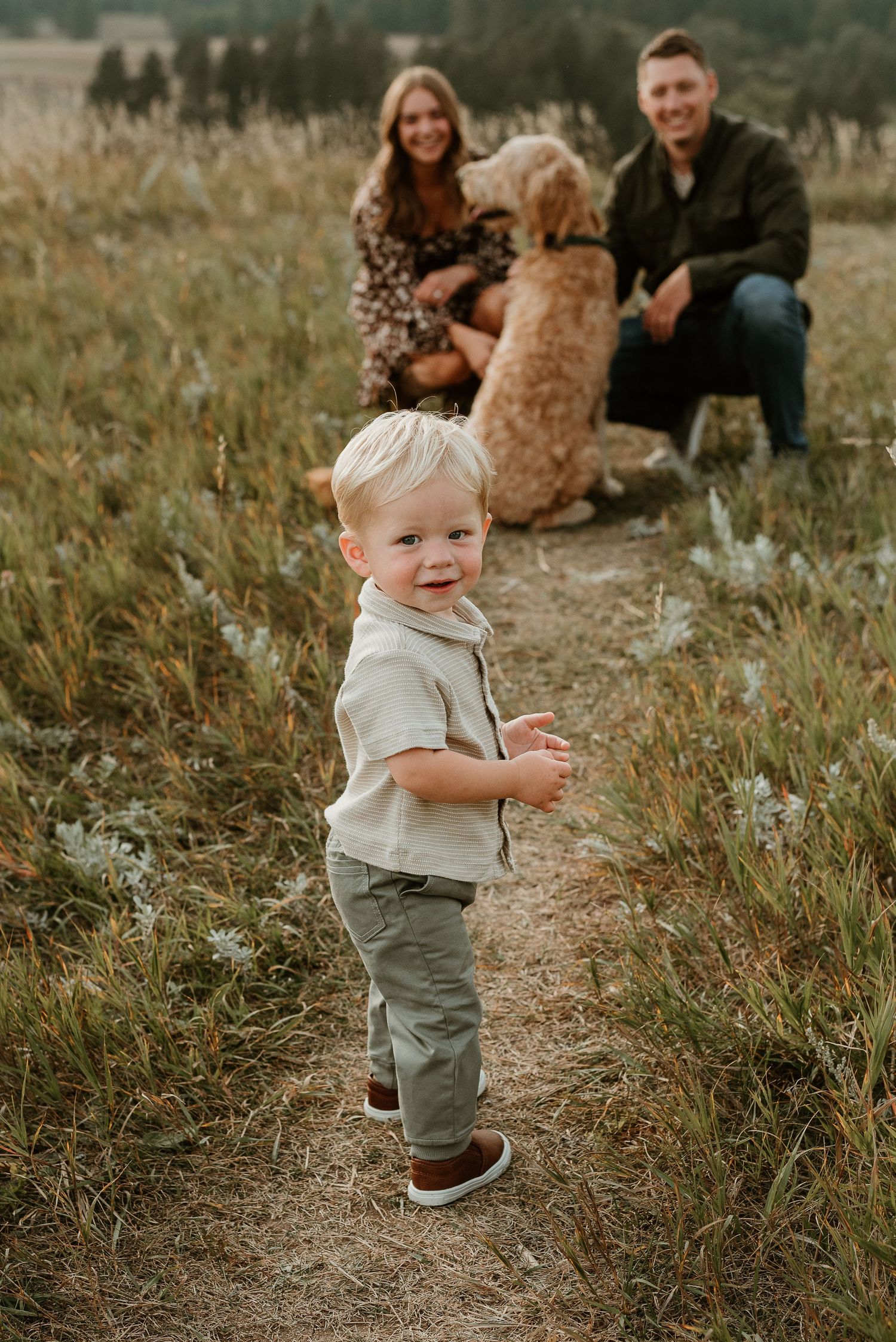 A toddler walks on a dirt path wearing sage green overalls and brown shoes in a natural outdoor setting.