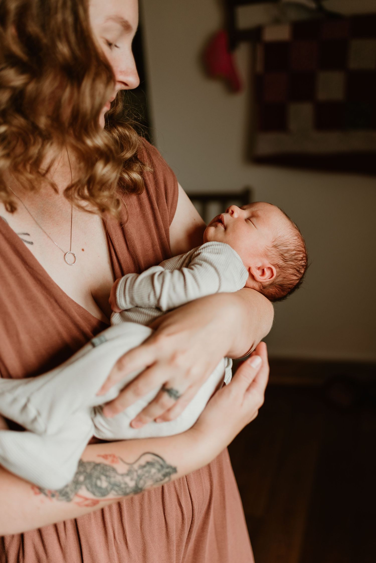 A parent in a dusty rose colored shirt tenderly holds and comforts a newborn baby wrapped in white.