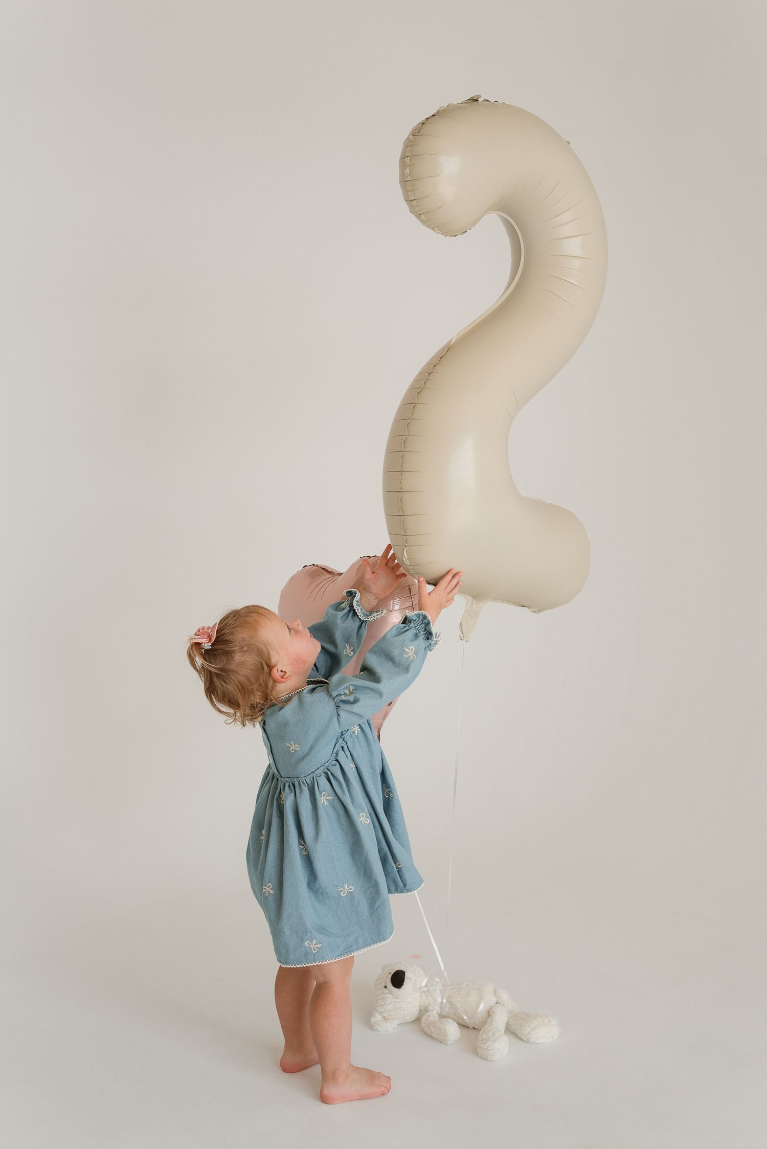 Toddler in blue dress reaching up to touch large number 2 balloon against white backdrop.