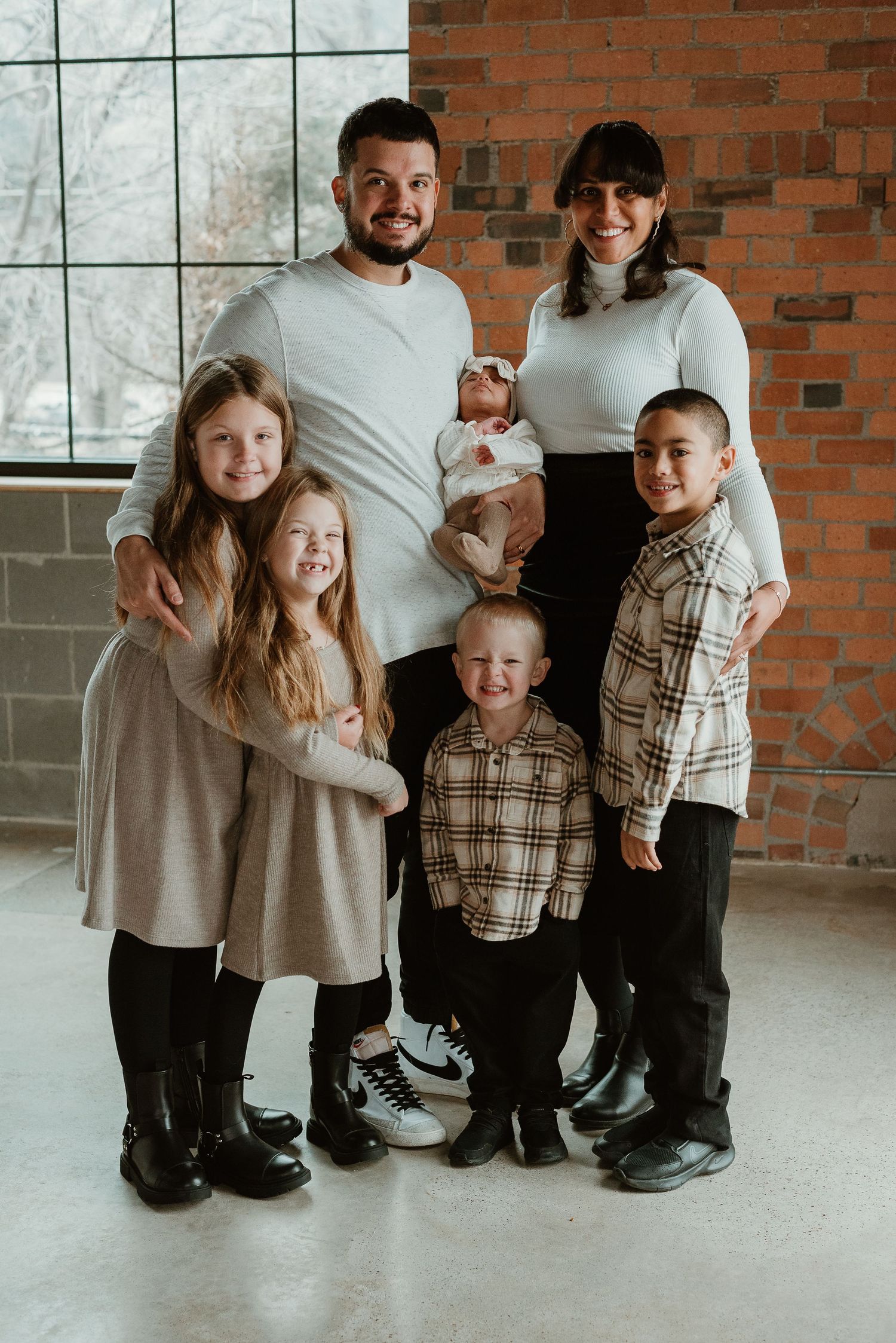 Family of six poses together in coordinated cream and plaid outfits against brick wall backdrop.