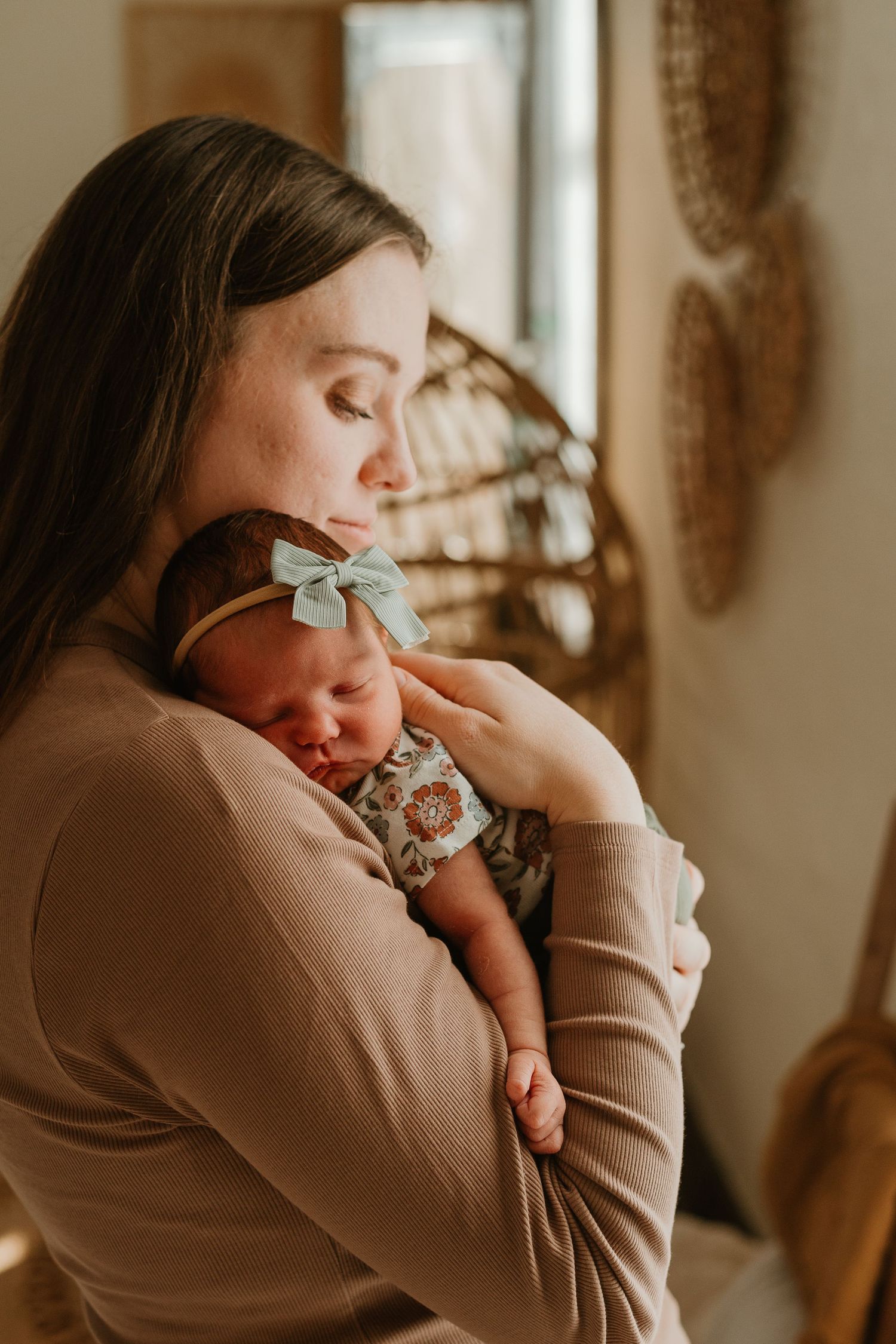 A person in a beige sweater holds a baby while sitting in a warmly lit room with natural window light.