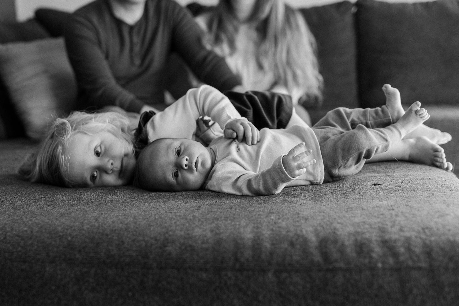 Two young children lying together on a gray couch in a sweet black and white photograph.