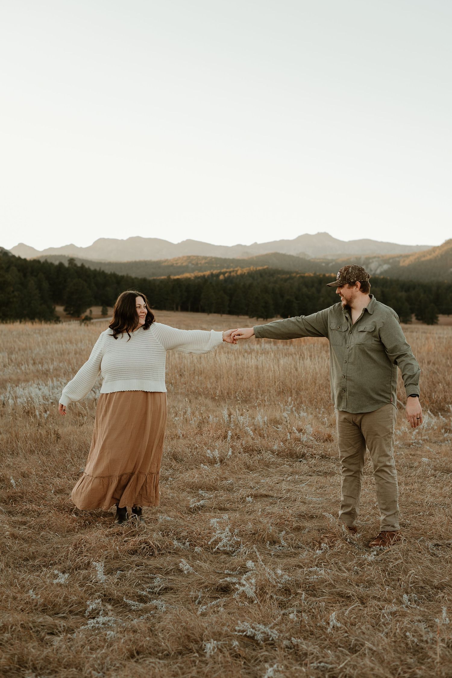 A couple dances playfully in a golden field at sunset with mountains in the background.