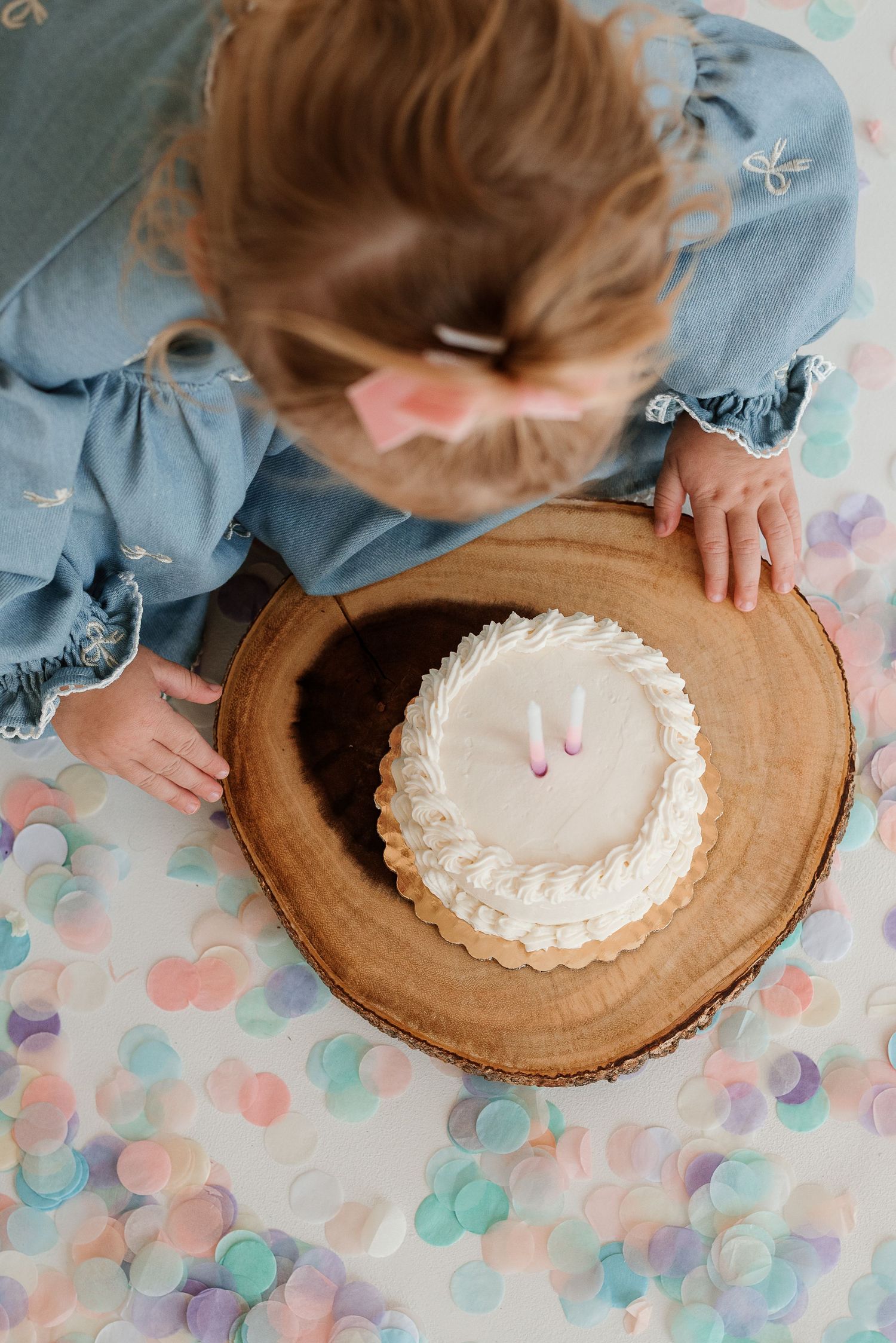 A person decorates a small white cake placed on a rustic wooden board surrounded by pastel colored confetti.