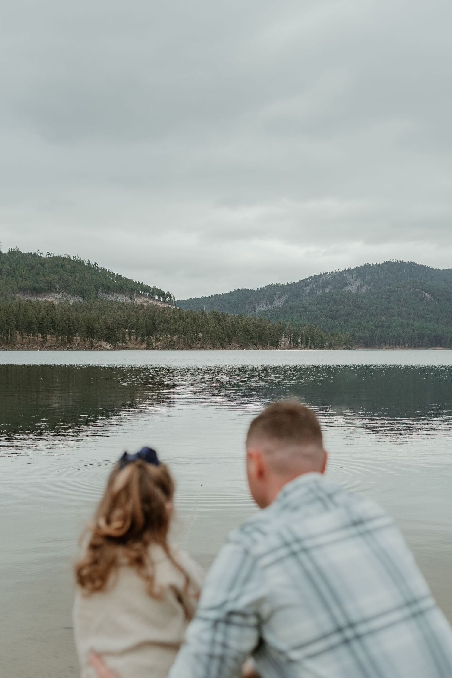 Two people sit by a peaceful mountain lake on an overcast day.