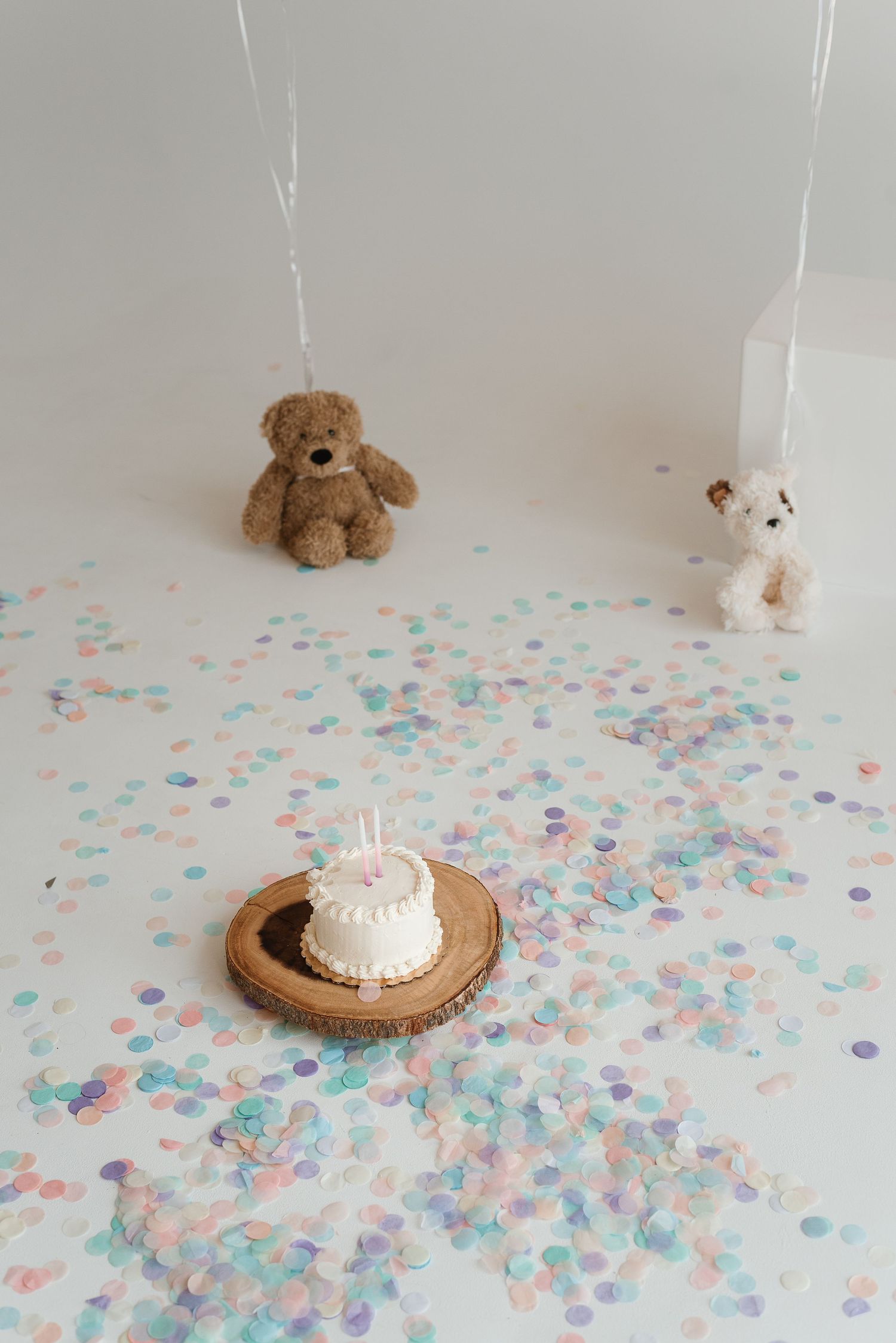 A minimalist birthday setup with pastel confetti, stuffed animals, and a white cake on a wooden plate.