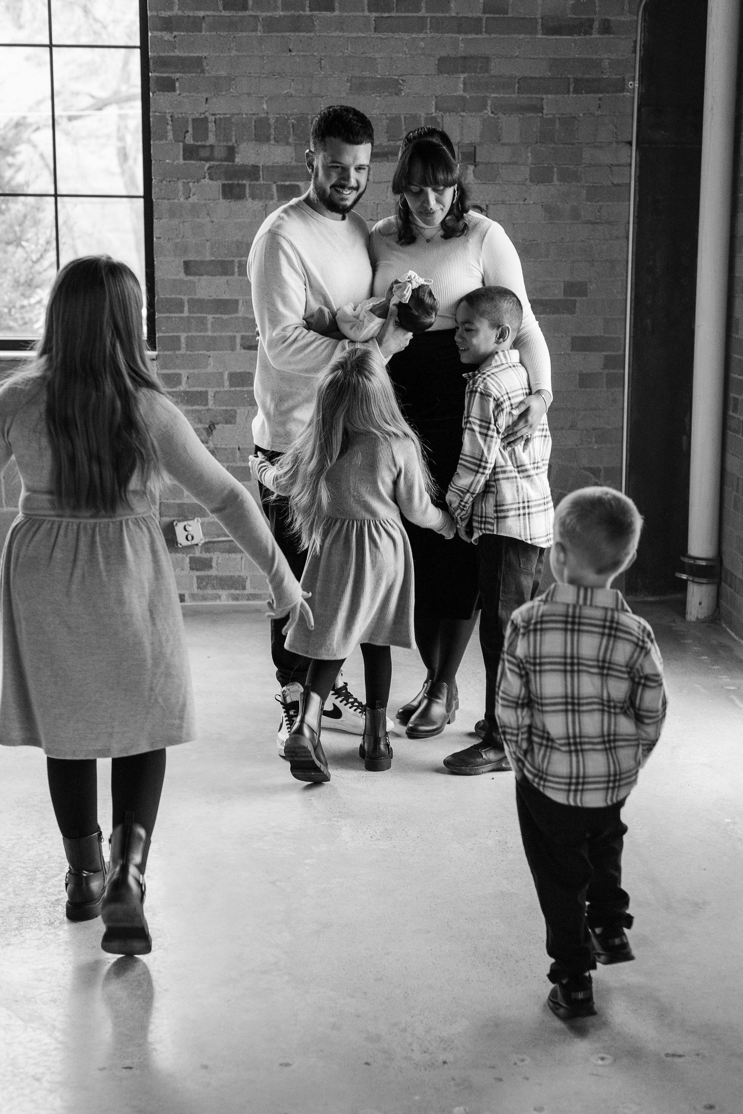 Black and white photo of family gathered together during indoor photoshoot against brick wall.