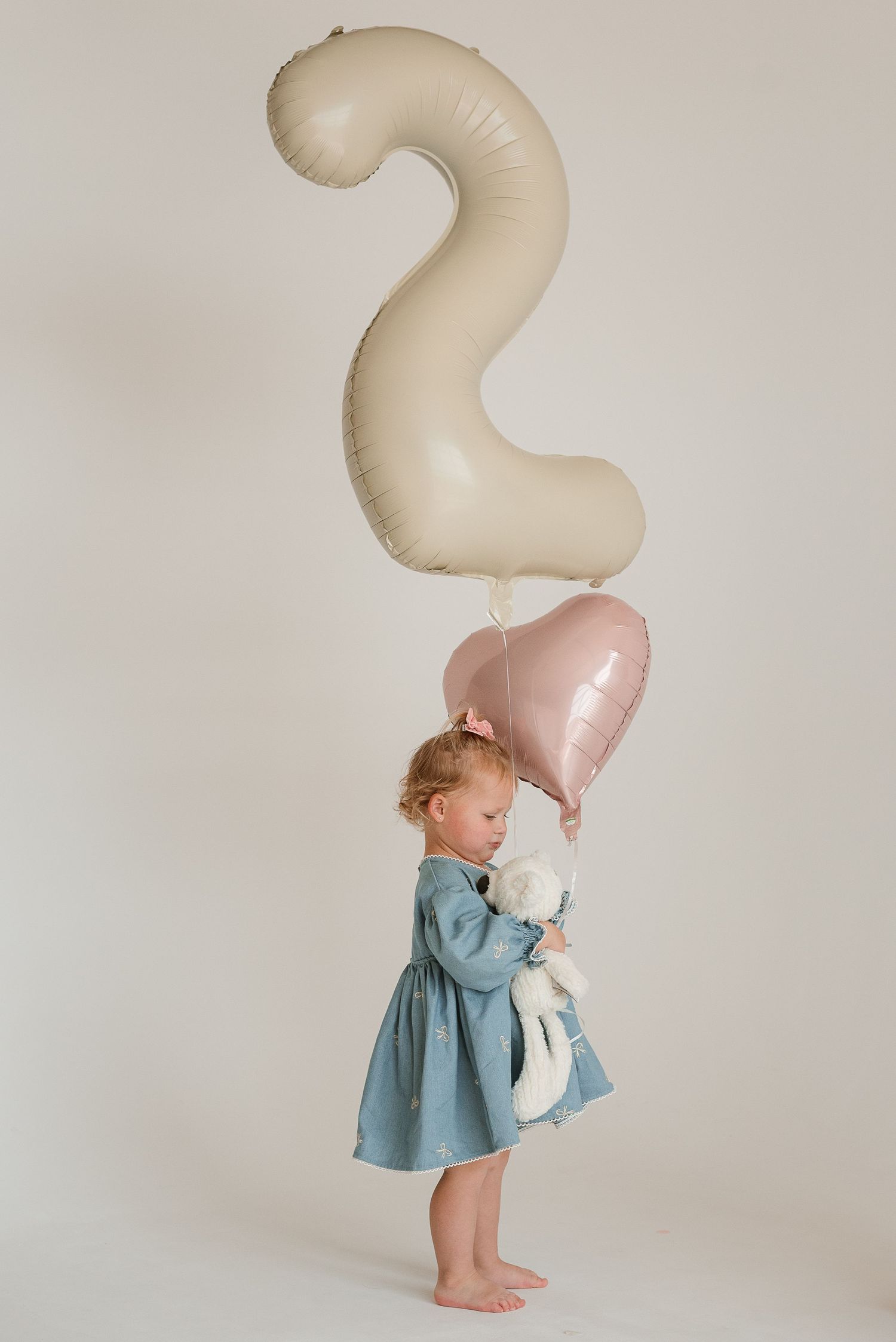 A toddler in blue dress plays with a large number 2 balloon against white backdrop.