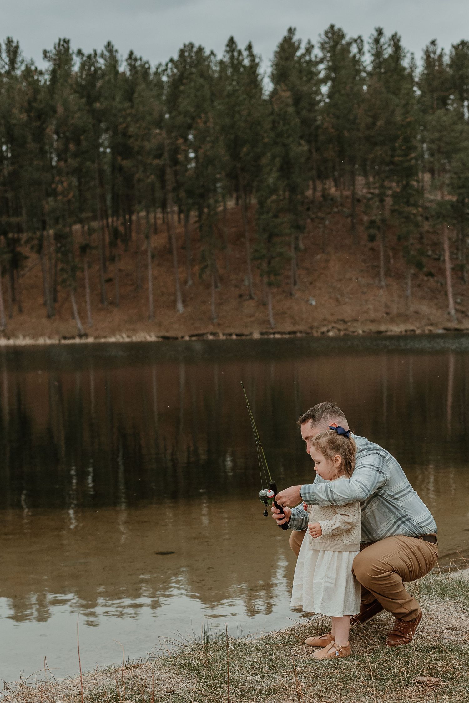 A parent and child share a fishing moment by a serene lake surrounded by pine trees.
