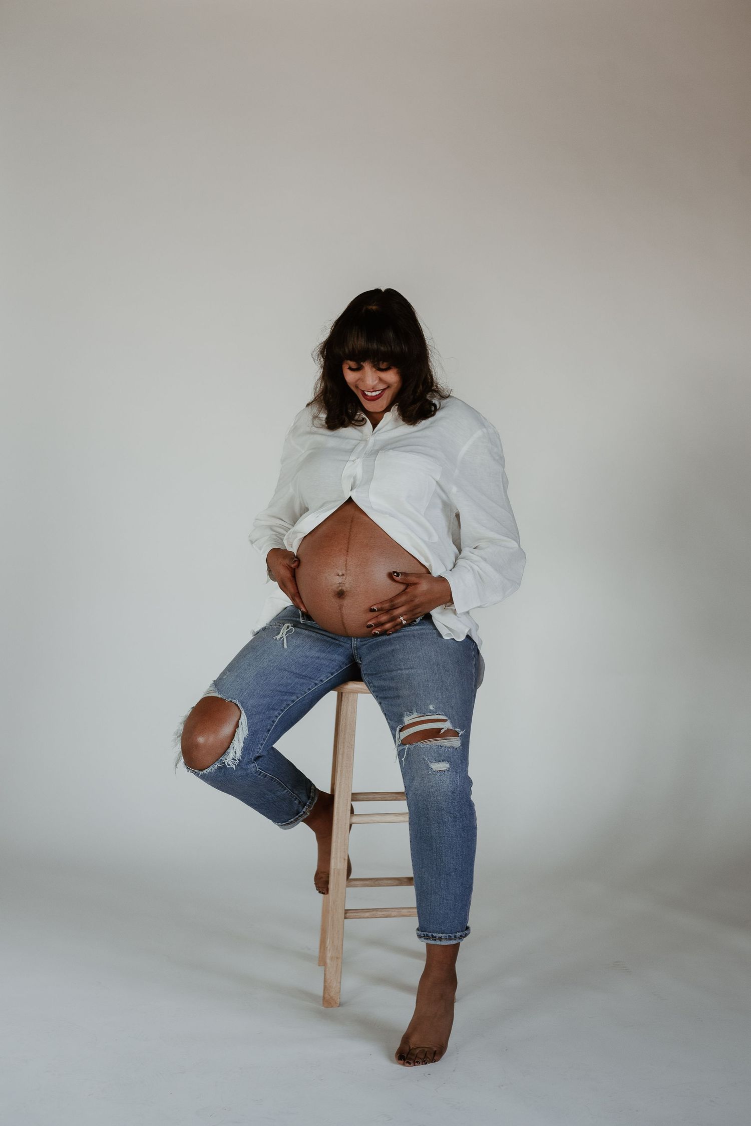 Expectant mother in white shirt and ripped jeans poses casually on wooden stool.
