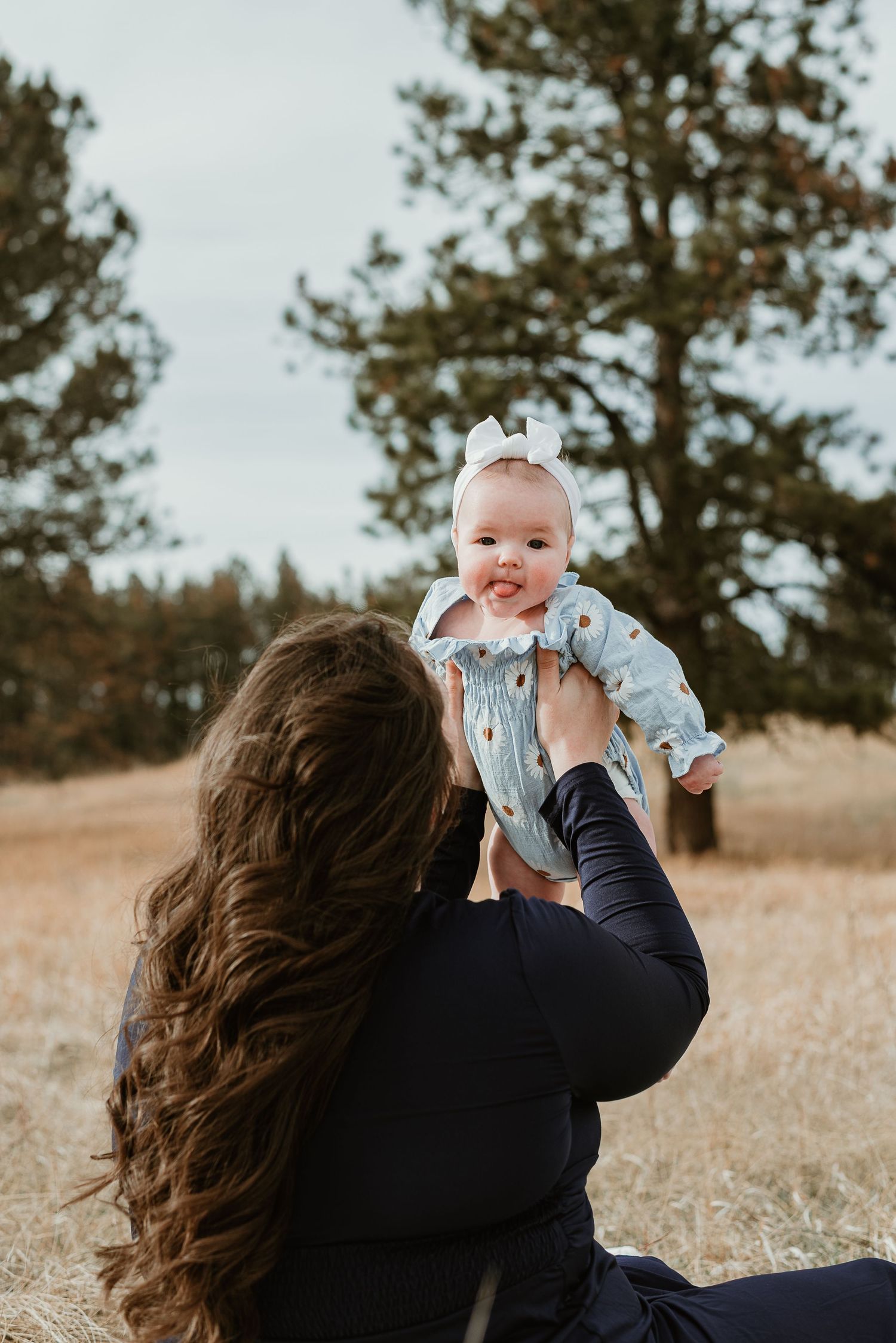 Baby in light blue dress being held up against a natural outdoor backdrop.