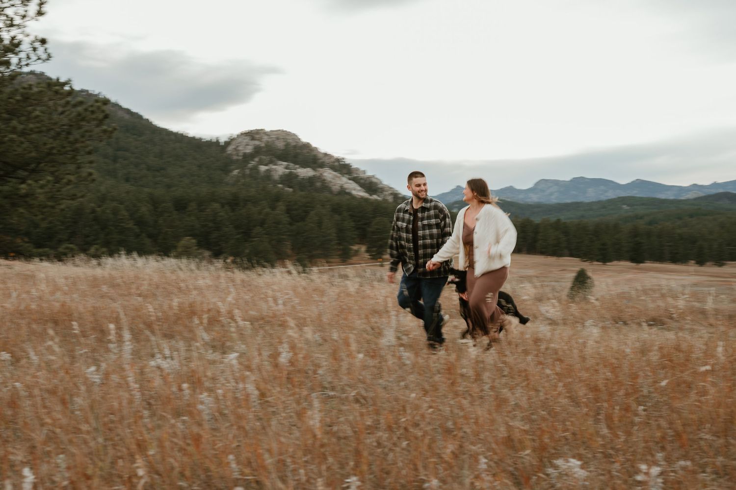 A couple walking through a golden meadow with mountains and forest in the background on an overcast day.