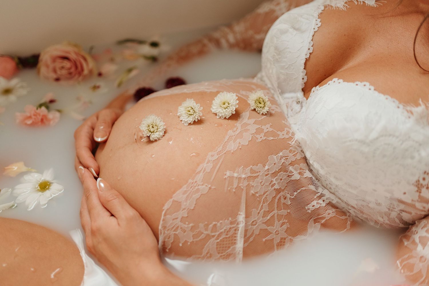 Delicate white lace covers a pregnant belly with small white flowers scattered on a soft pink background.