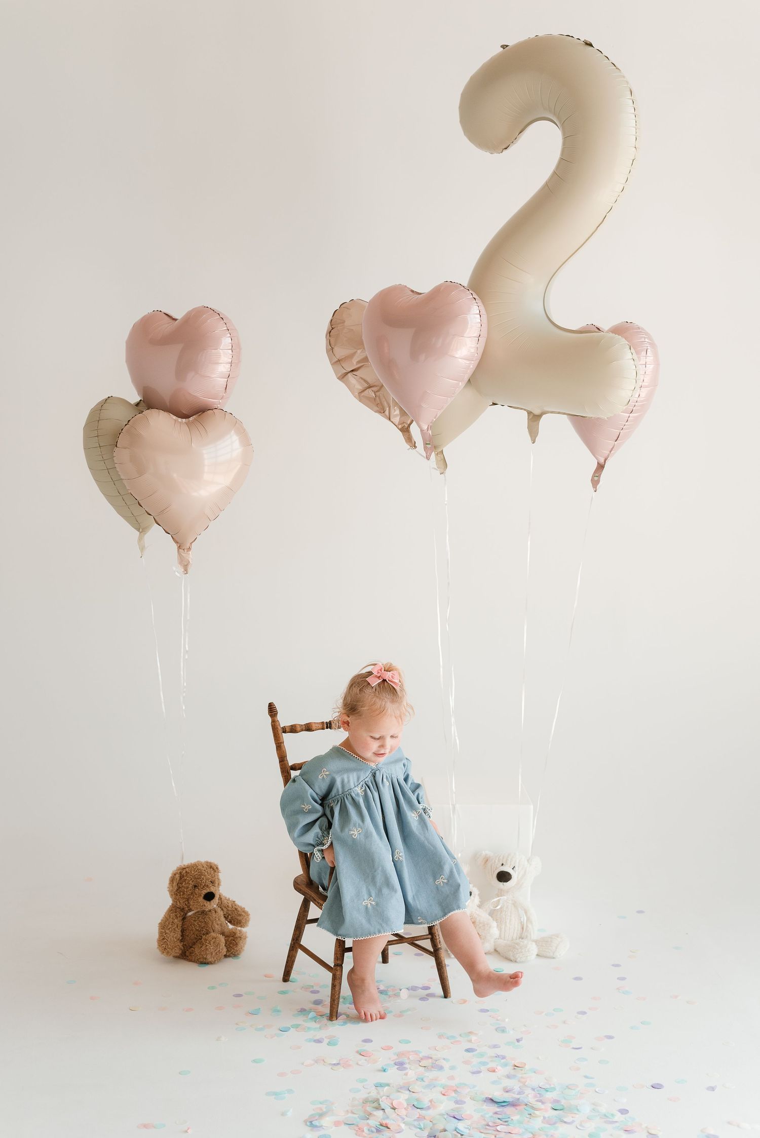 A birthday celebration setup with pink heart balloons, a small cake, and a teddy bear on a wooden chair against white backdrop.