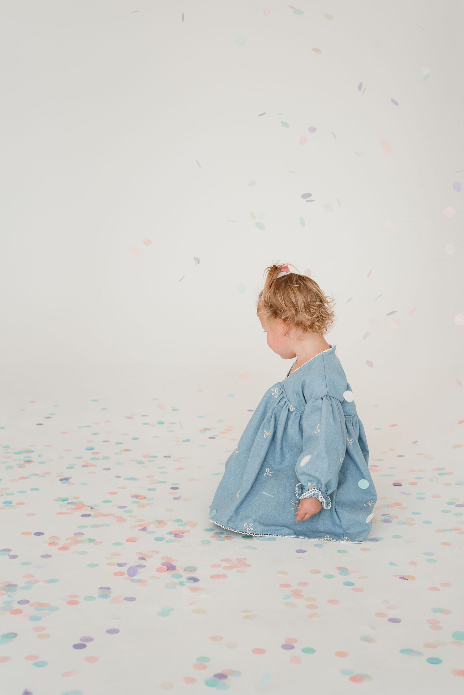 A small child in a blue dress sits on a white floor scattered with pink and blue confetti dots.