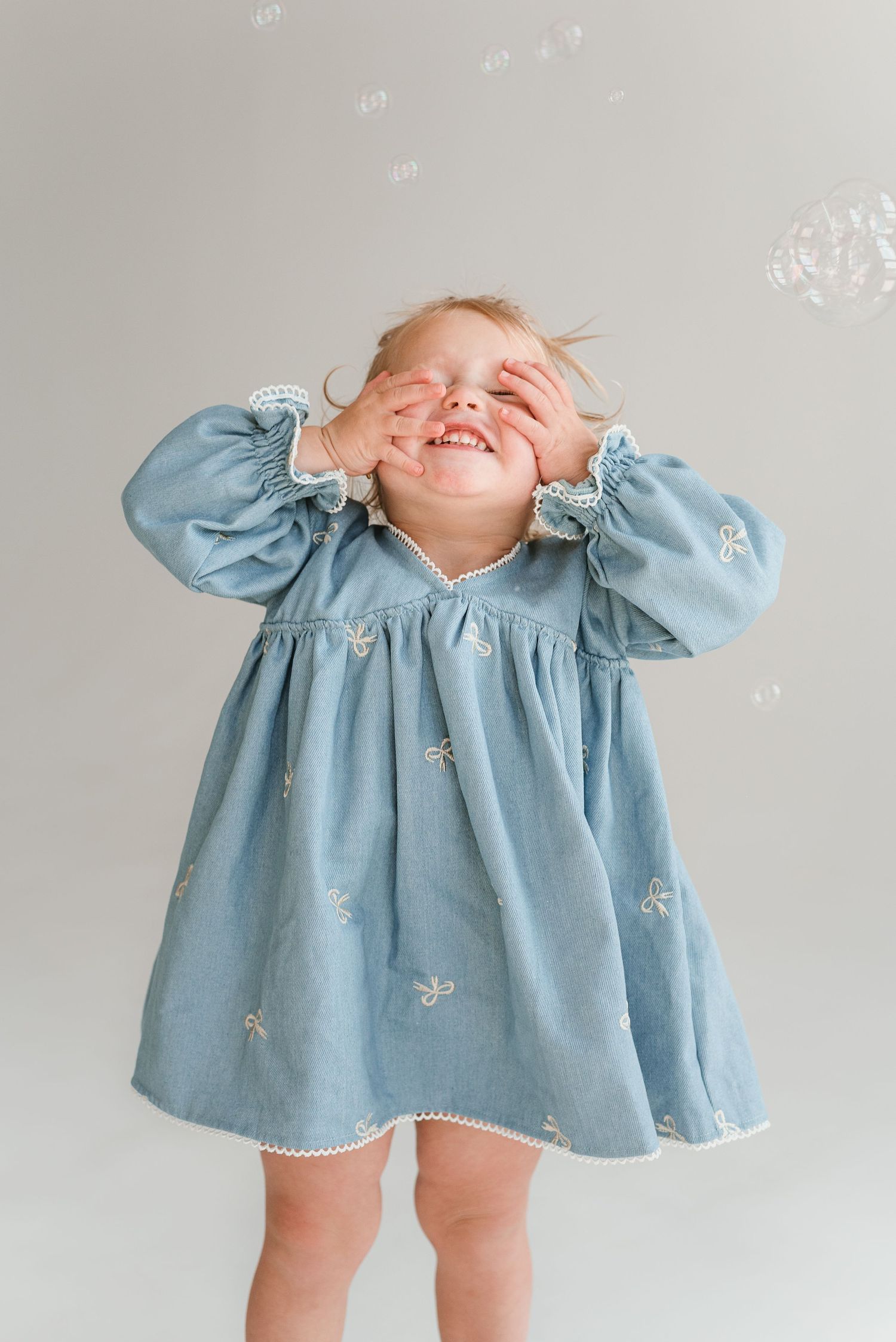 A small child in a light blue dress plays and giggles while covering their face in a series of candid photos.