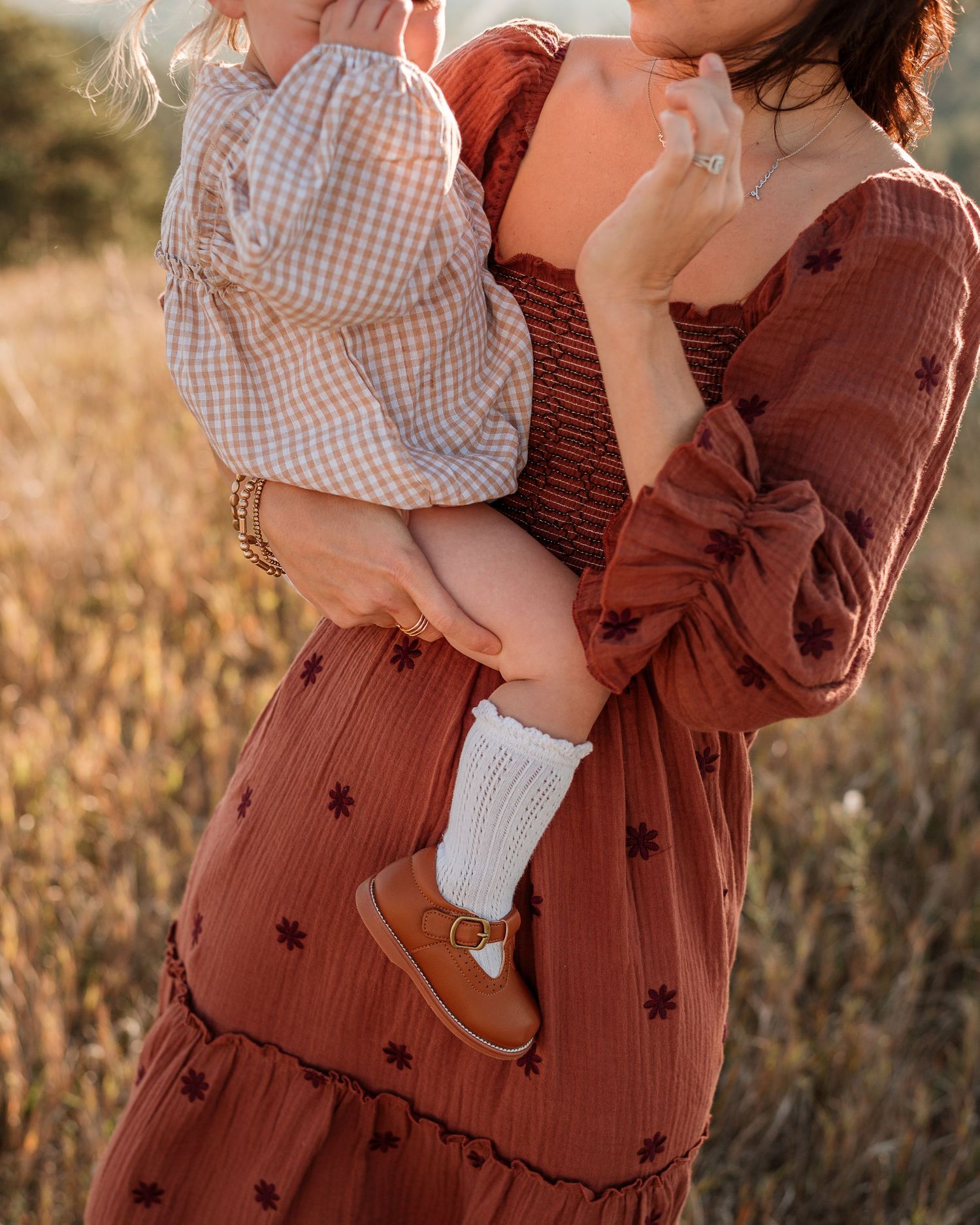 A person wearing a brown sweater with stars holds a small child in a gingham dress during a golden hour photoshoot.