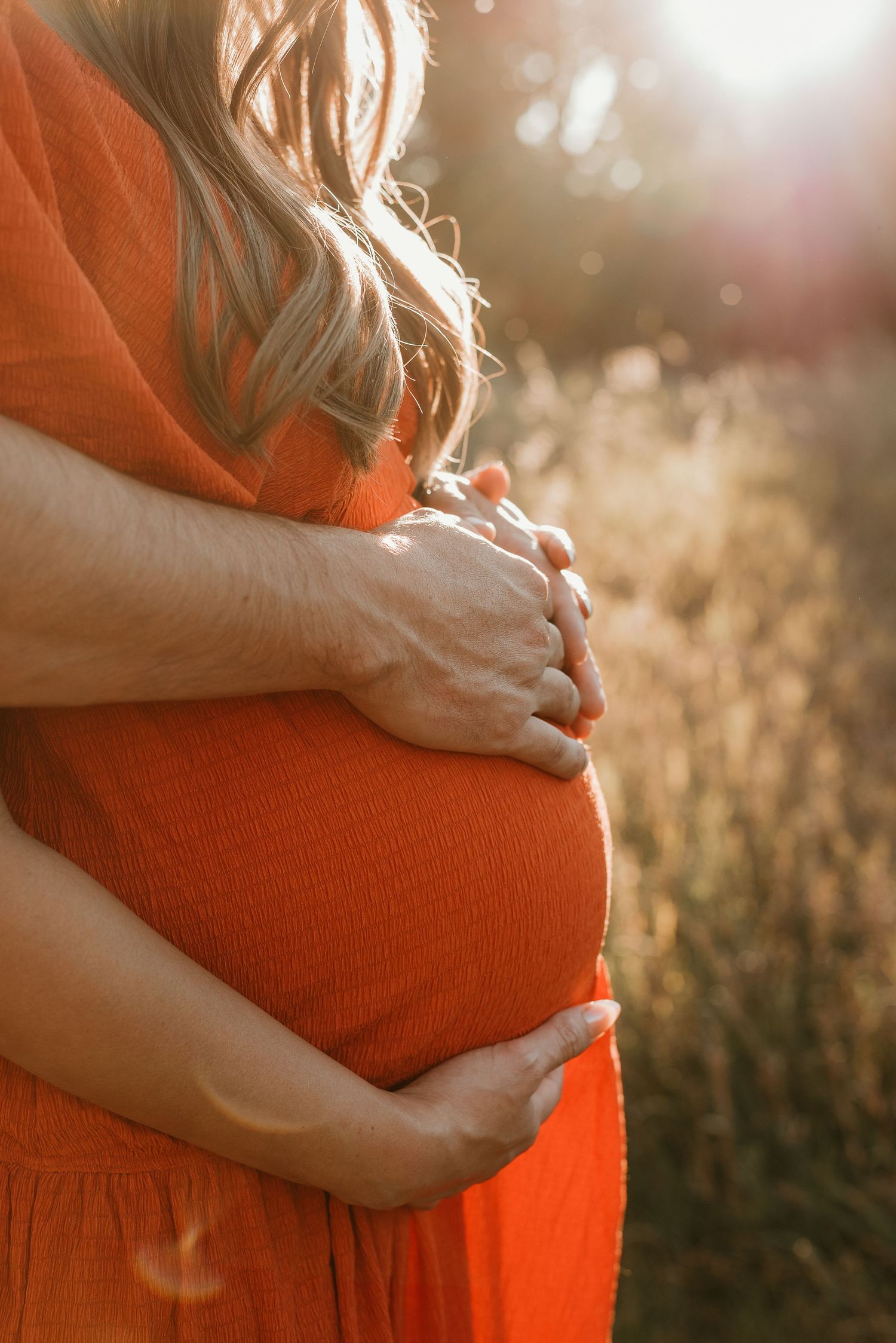 Close-up shot of hands cradling a pregnant belly in an orange dress with sunlit bokeh background.