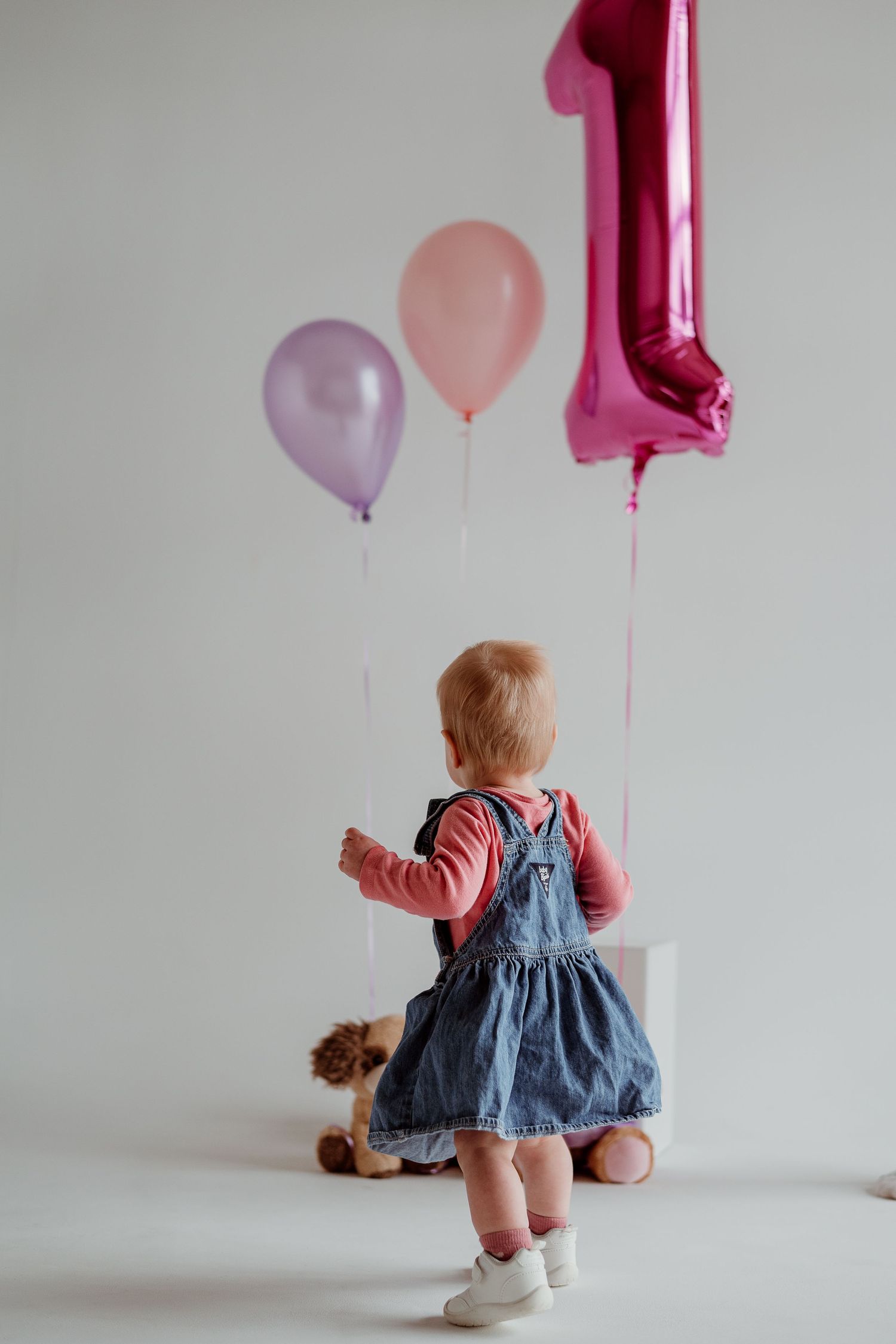 A small child in a denim dress and pink top walks under pink and purple birthday balloons against a white wall.