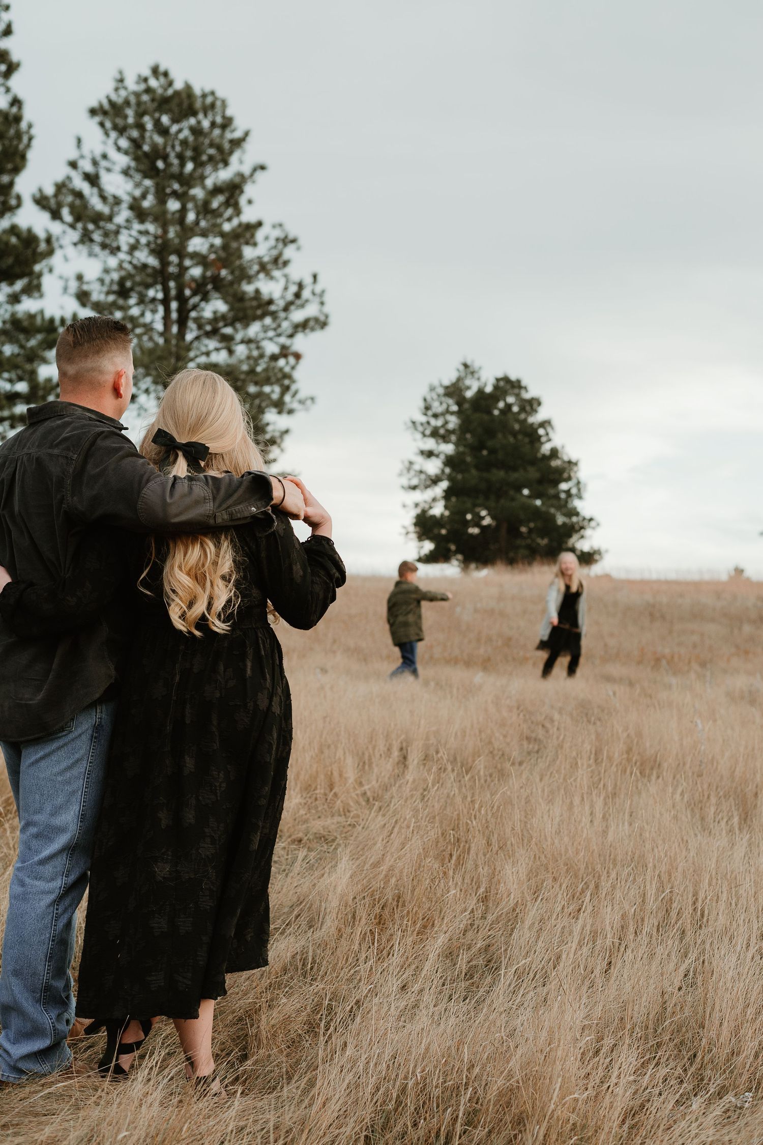 A couple embracing in a field while others can be seen in the distance on a cloudy day.