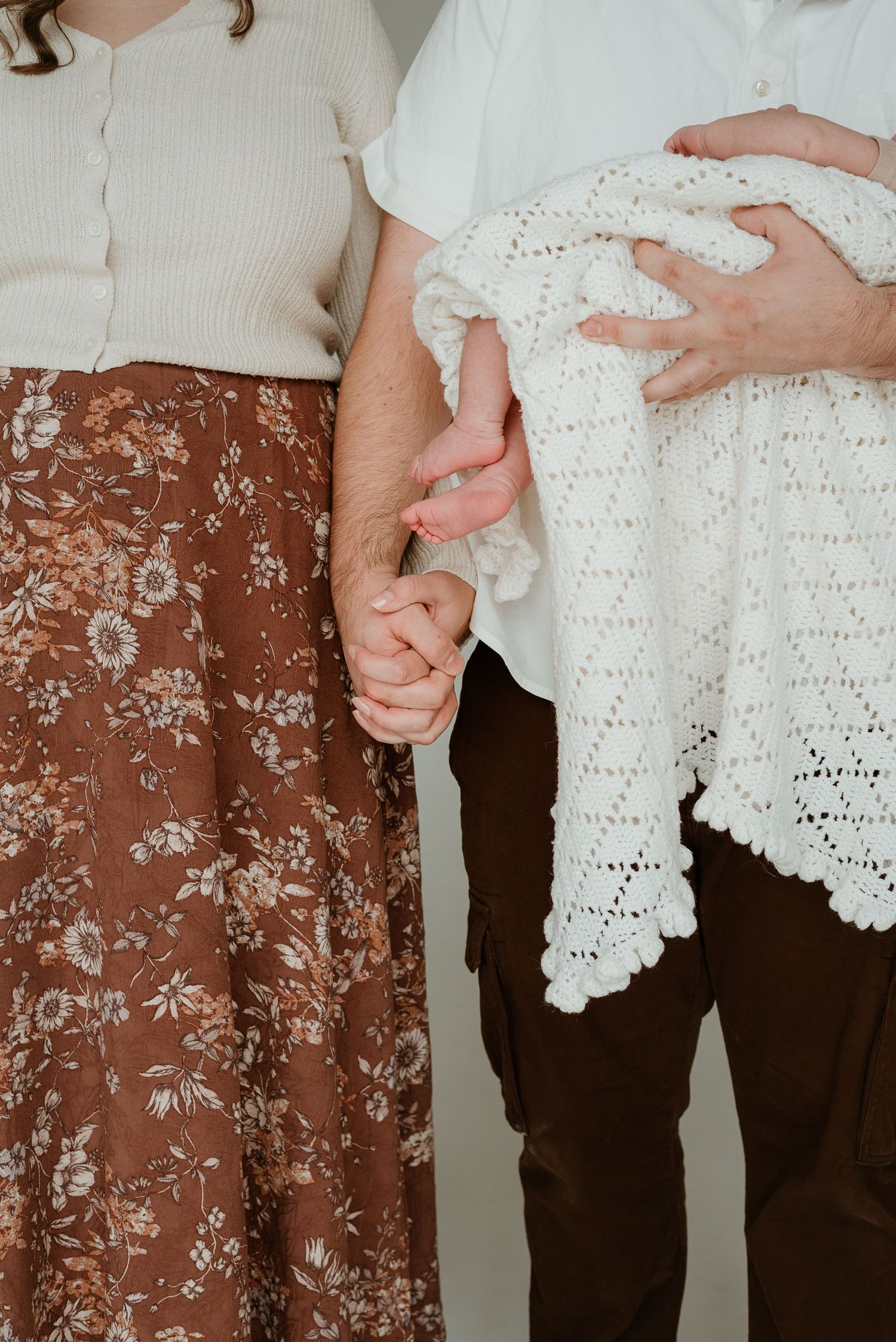 Close-up of hands holding together while one person wears floral pants and another holds a white crocheted blanket.