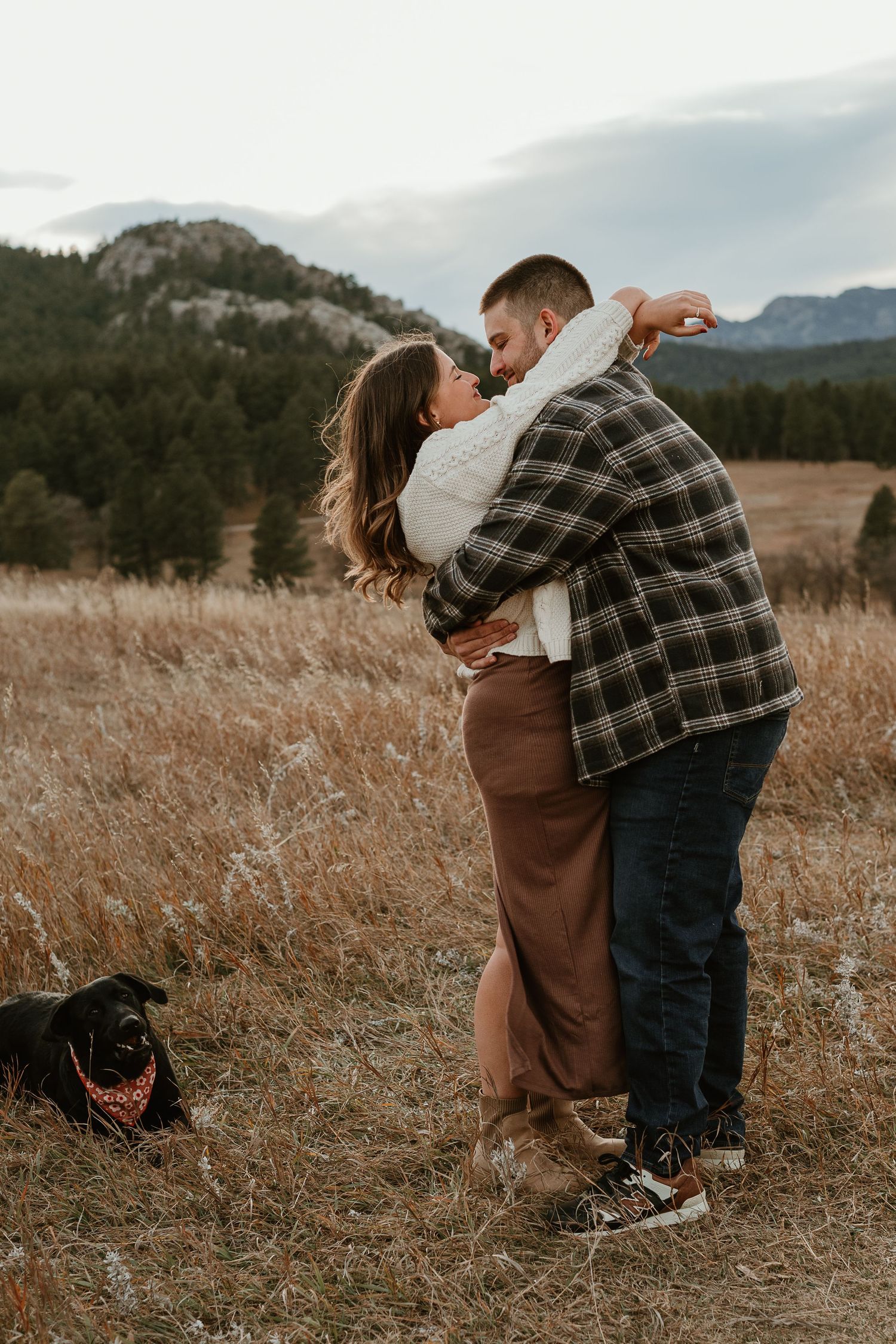 A couple embraces in a romantic moment on a mountainside field at sunset while their dog watches nearby.