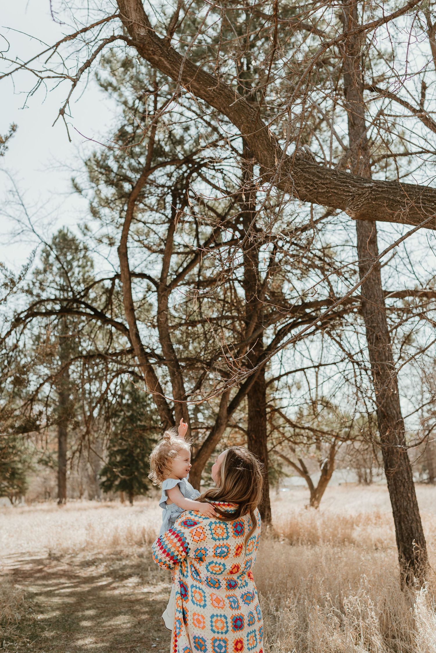 People in light clothing and a colorful cardigan enjoy a moment among tall pine trees.