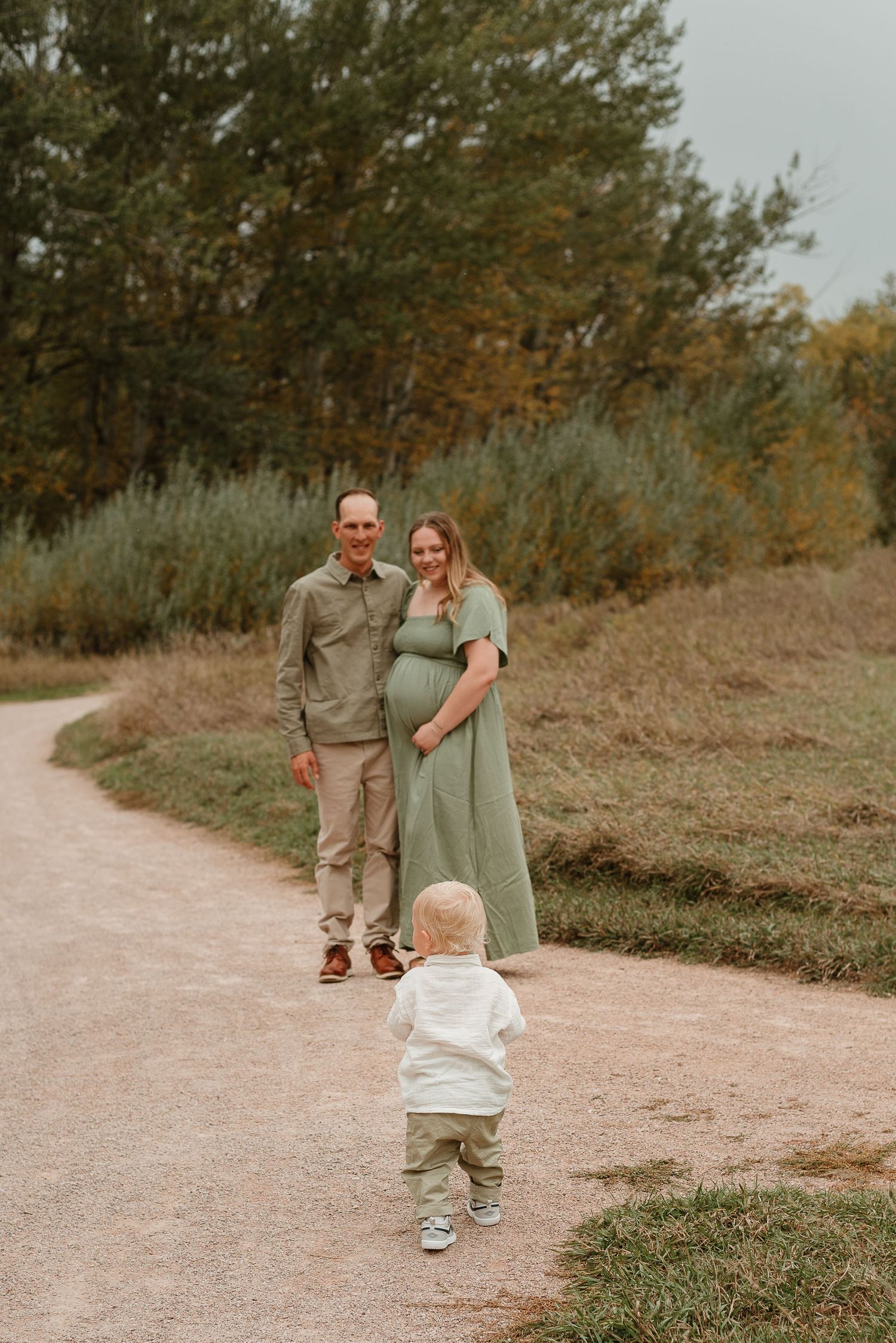 A toddler in white walks on a dirt path towards two people standing together in a natural outdoor setting.