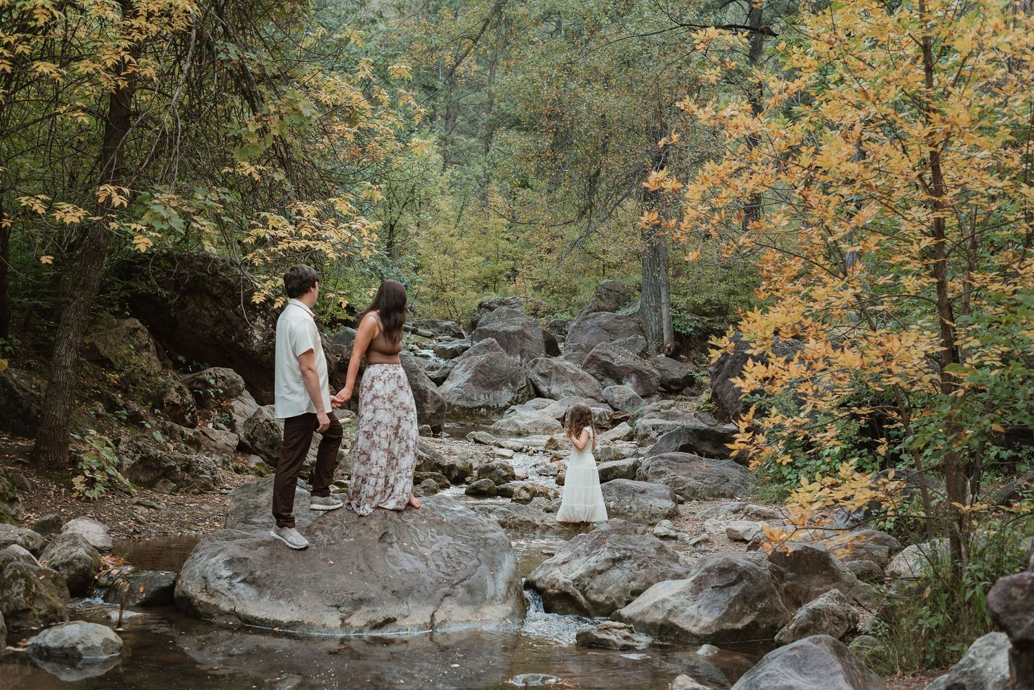 A couple poses for wedding photos along a rocky stream surrounded by autumn foliage in a forest setting.