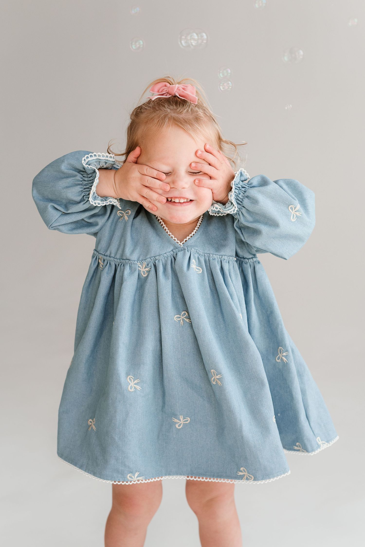Playful toddler in blue embroidered dress covers eyes while smiling and playing peekaboo against light background.
