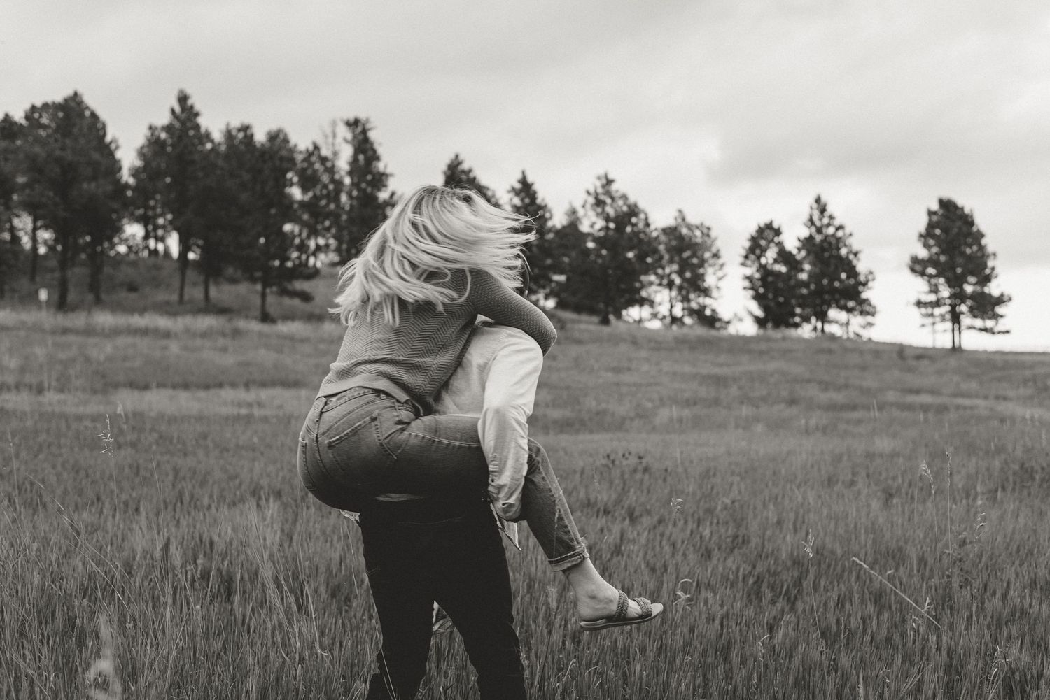 Black and white photo of a couple embracing in a grassy field with pine trees on the horizon.