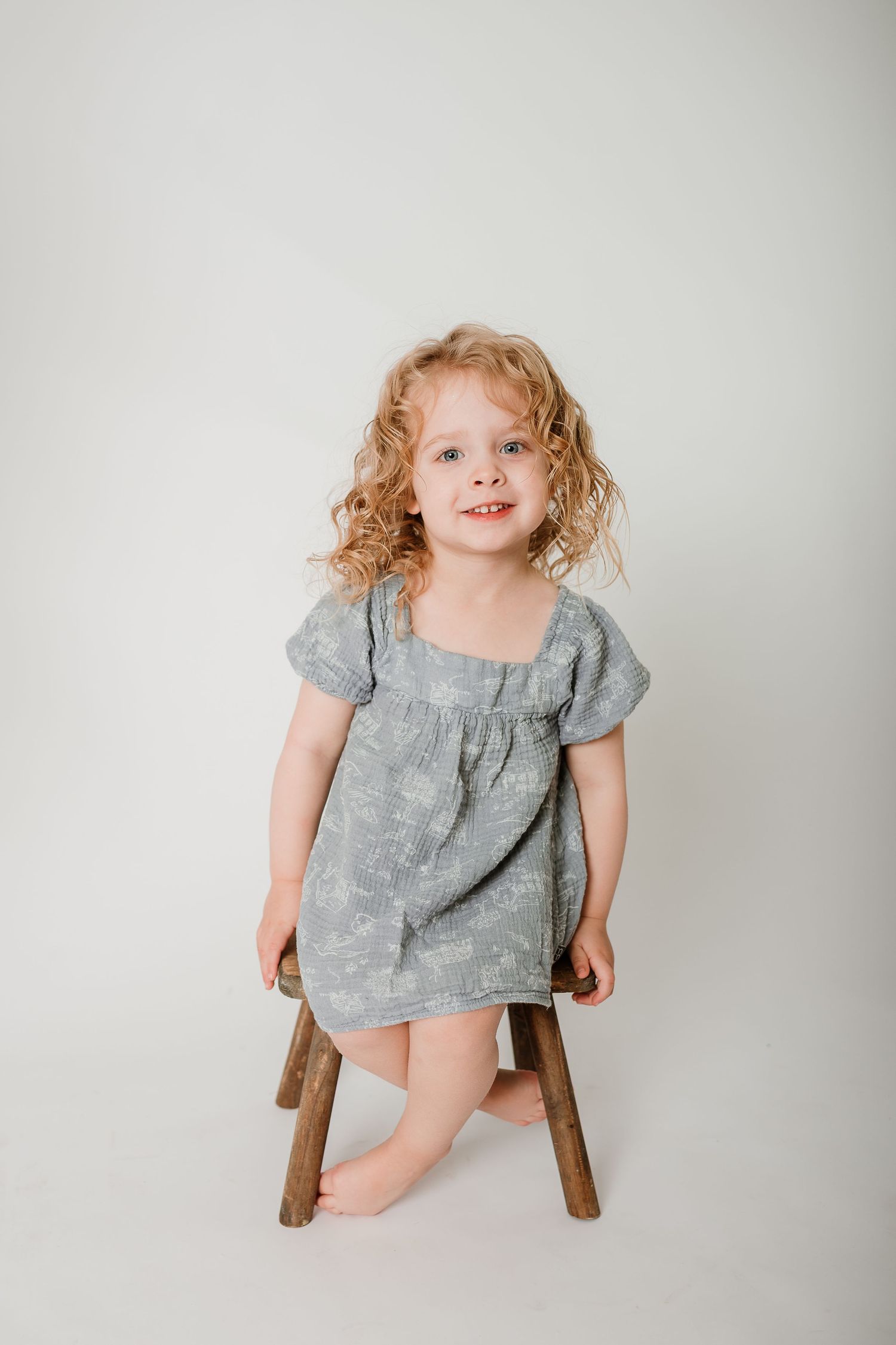 Child wearing a gray linen dress sits on a wooden stool against a white studio backdrop in three different poses.