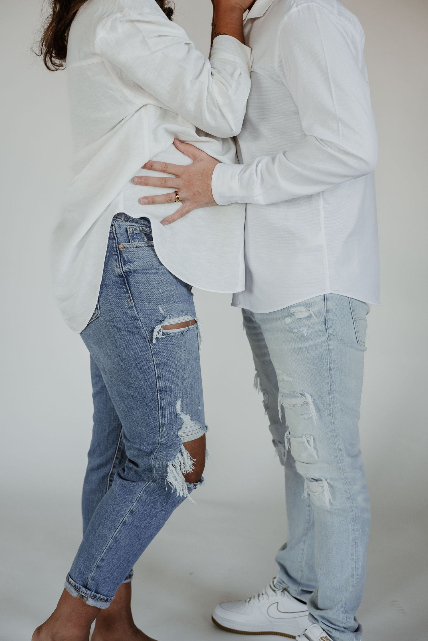 Couple in white shirts and distressed jeans embracing against white background.