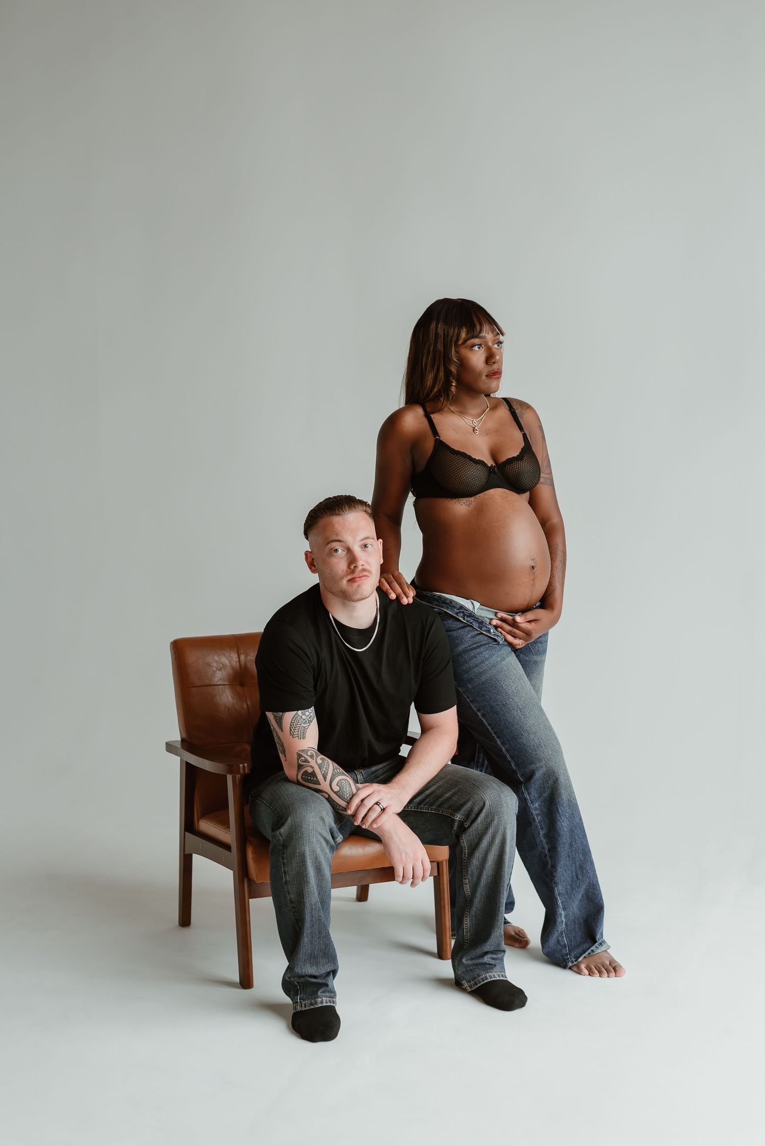 Two people sit casually on a wooden chair against a plain white backdrop in a minimalist studio setting.