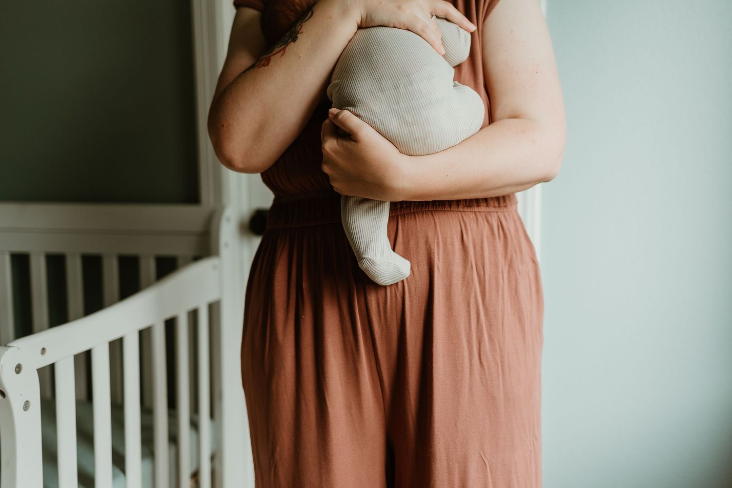 Pregnant woman in a flowing rust-colored dress cradles her baby bump while standing near a white crib.