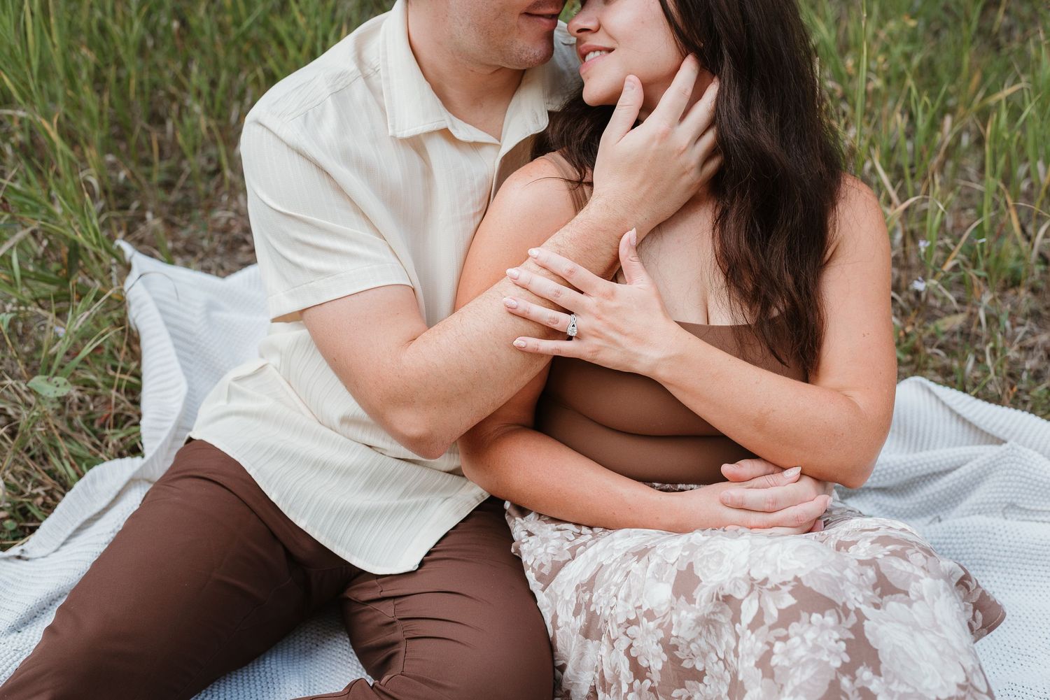 Romantic couple in white and brown outfits share tender moments while sitting together outdoors in a grassy field.