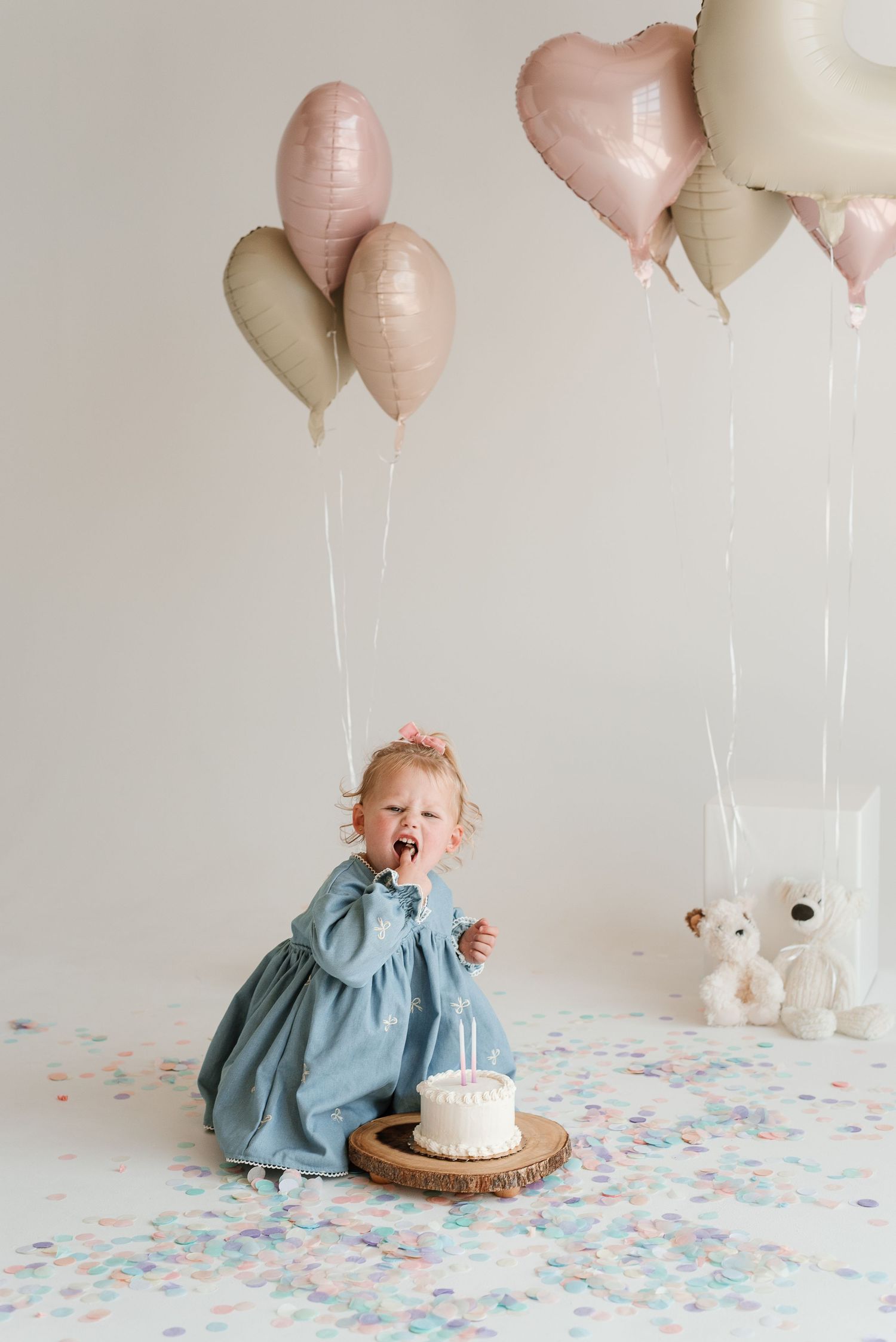 A festive birthday setup with pink and cream balloons, confetti, and a white cake on a wooden plate.