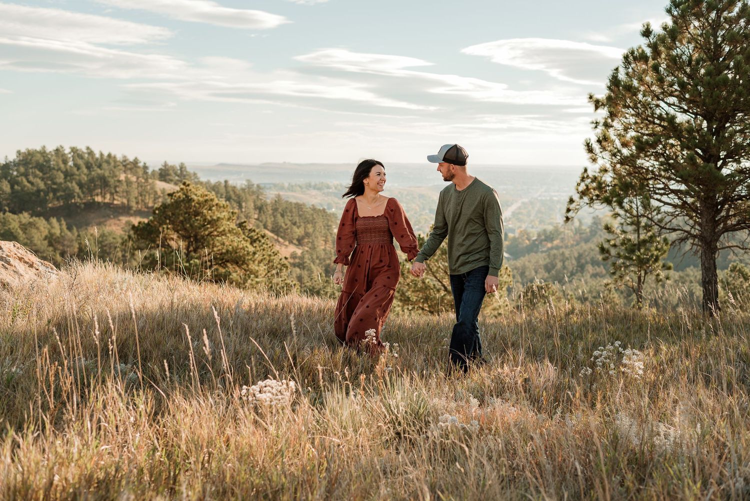A couple stands together in golden tall grass during sunset at a scenic mountain overlook.