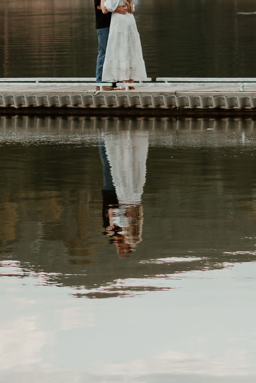 Reflection of a couple standing on a dock over calm lake waters.