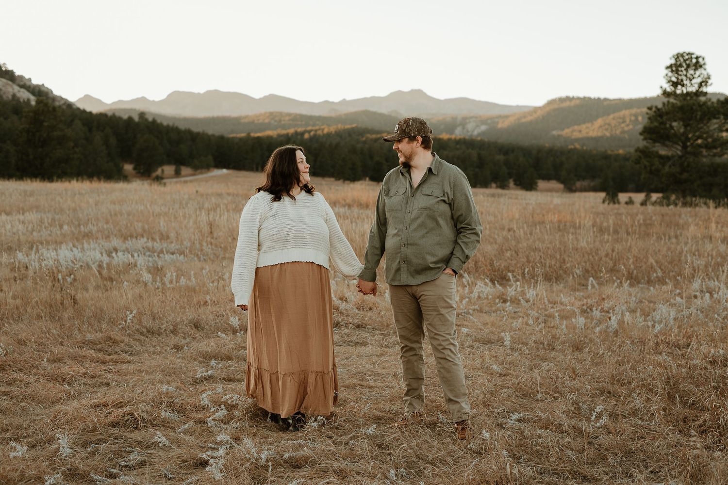 A couple holds hands while standing in a golden field with mountain ranges in the background at sunset.