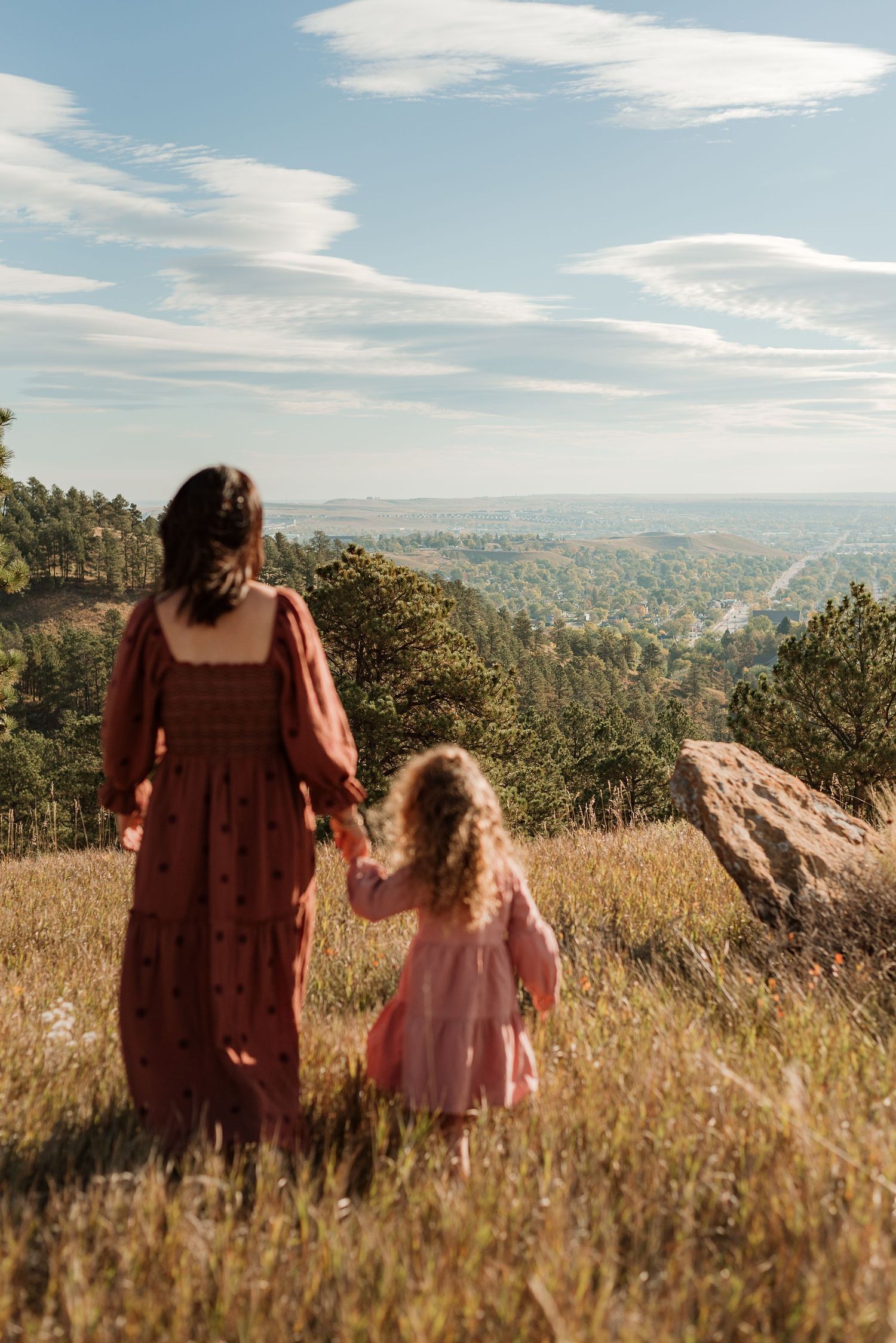Two people in dresses stand together in a golden meadow overlooking a scenic mountain landscape on a sunny day.