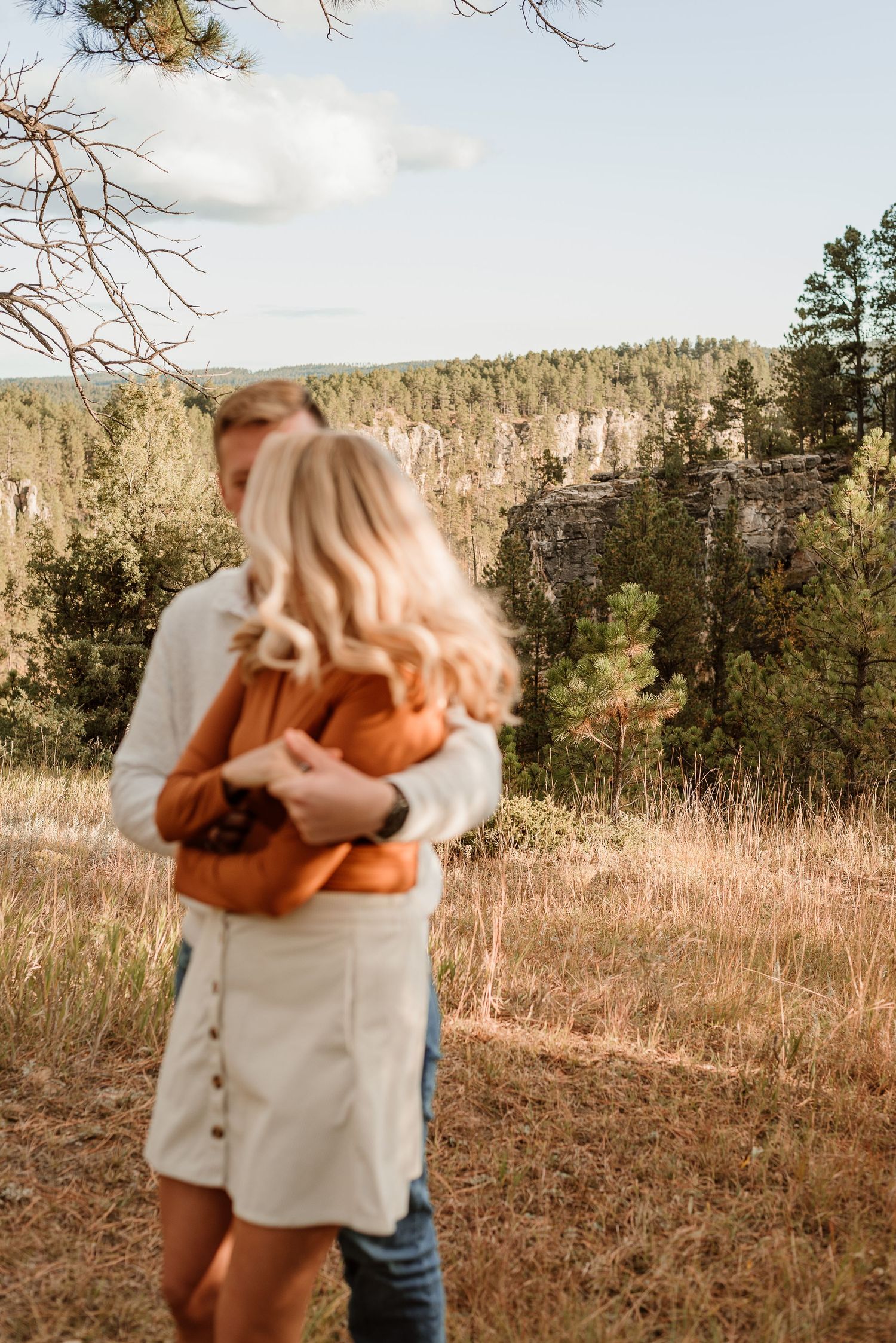 Couple embracing in a scenic mountain landscape with evergreen trees and golden grass at sunset.
