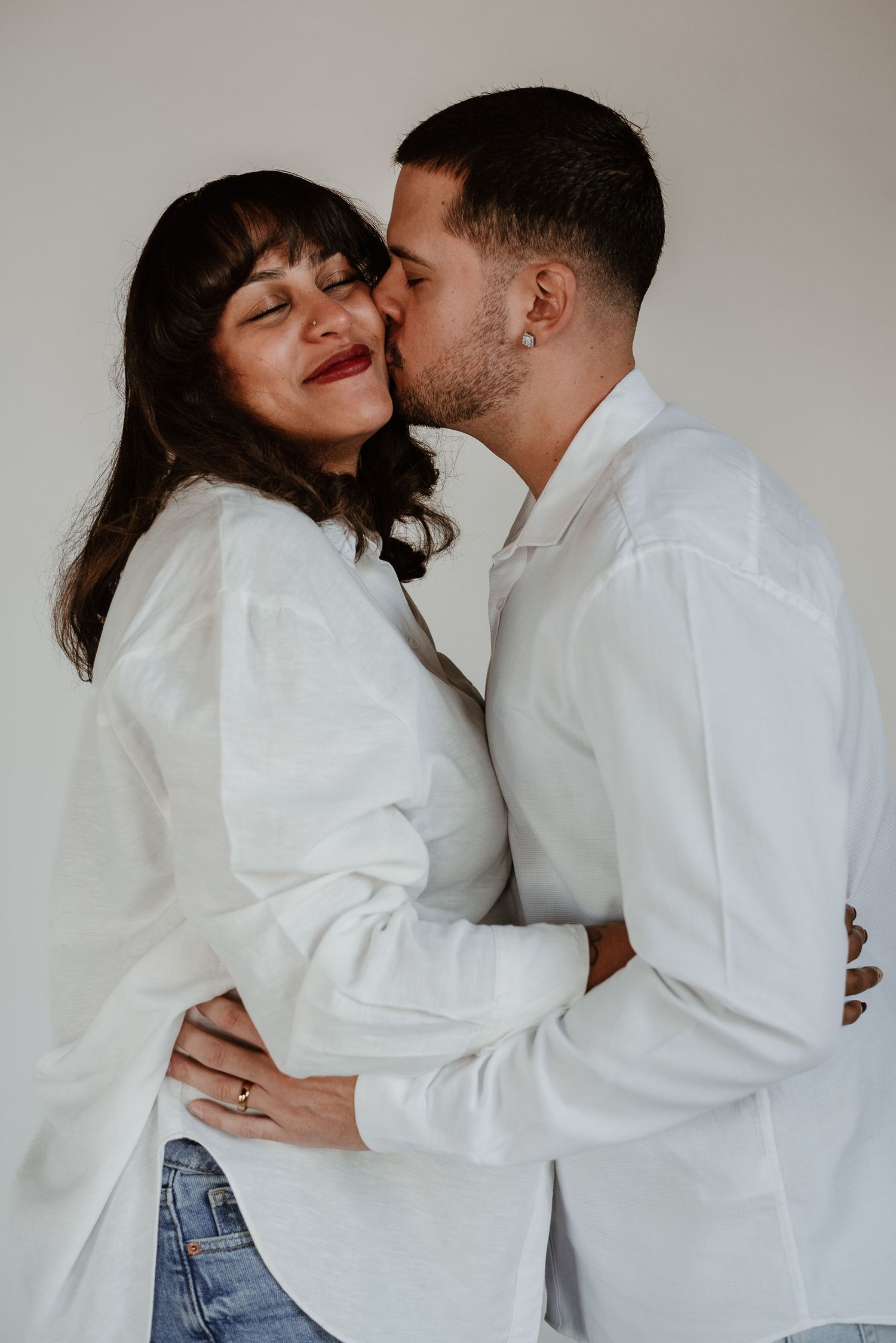 Couple in white shirts sharing intimate moment against neutral wall.