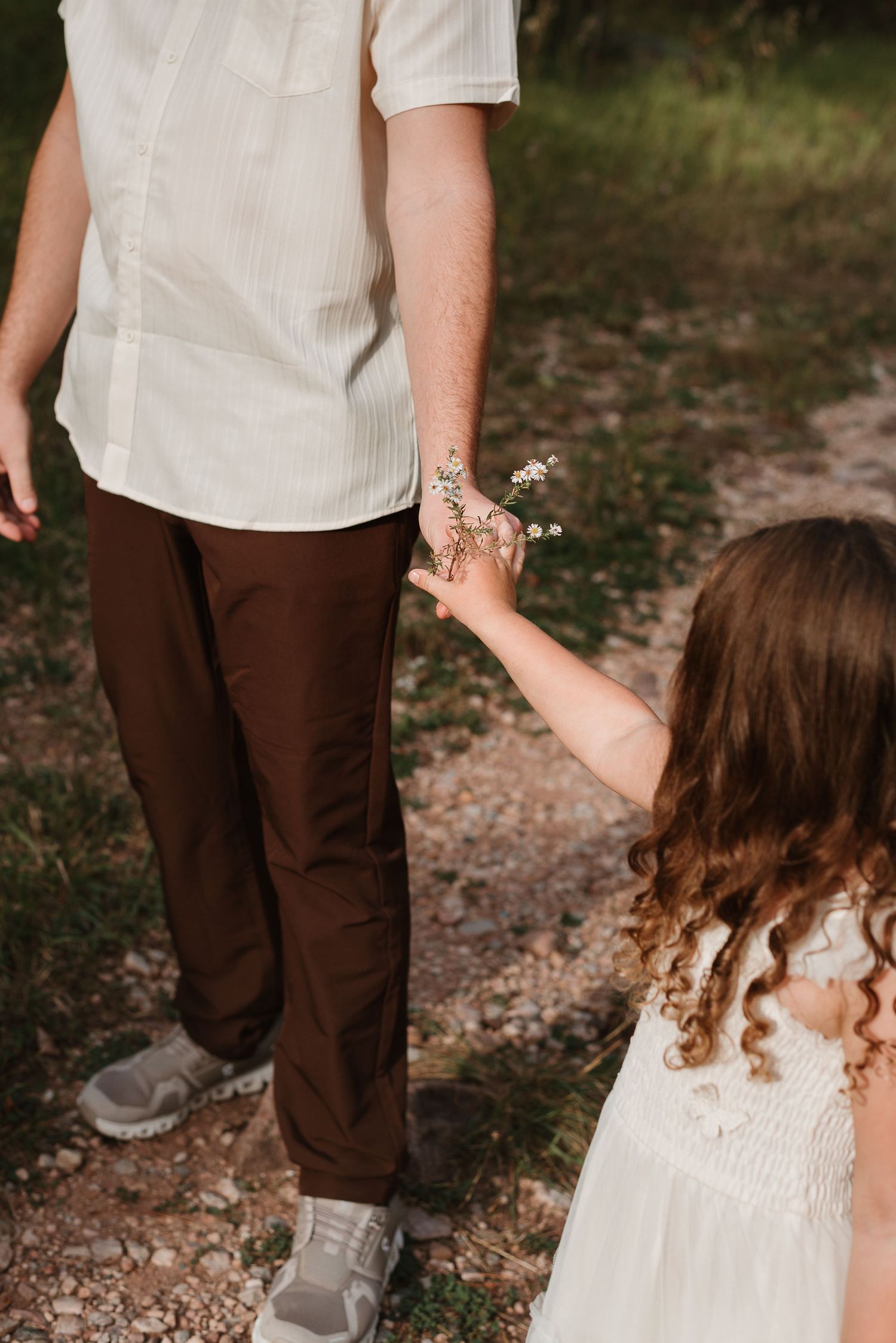 A small child in a white dress holds hands with someone wearing brown pants while walking on a gravel path.