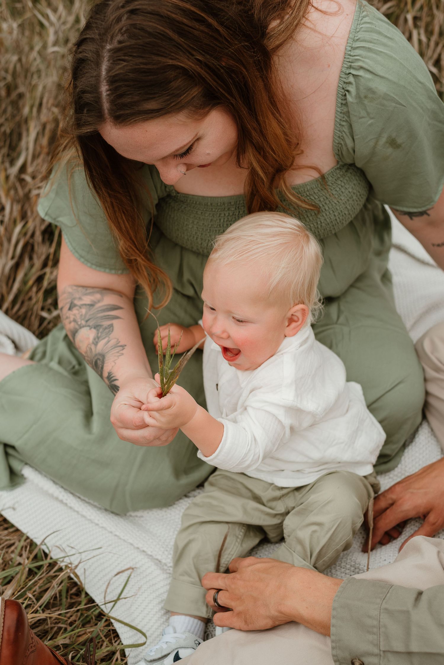 A tender moment between parent and child sitting in dry grass wearing sage green and white clothing.