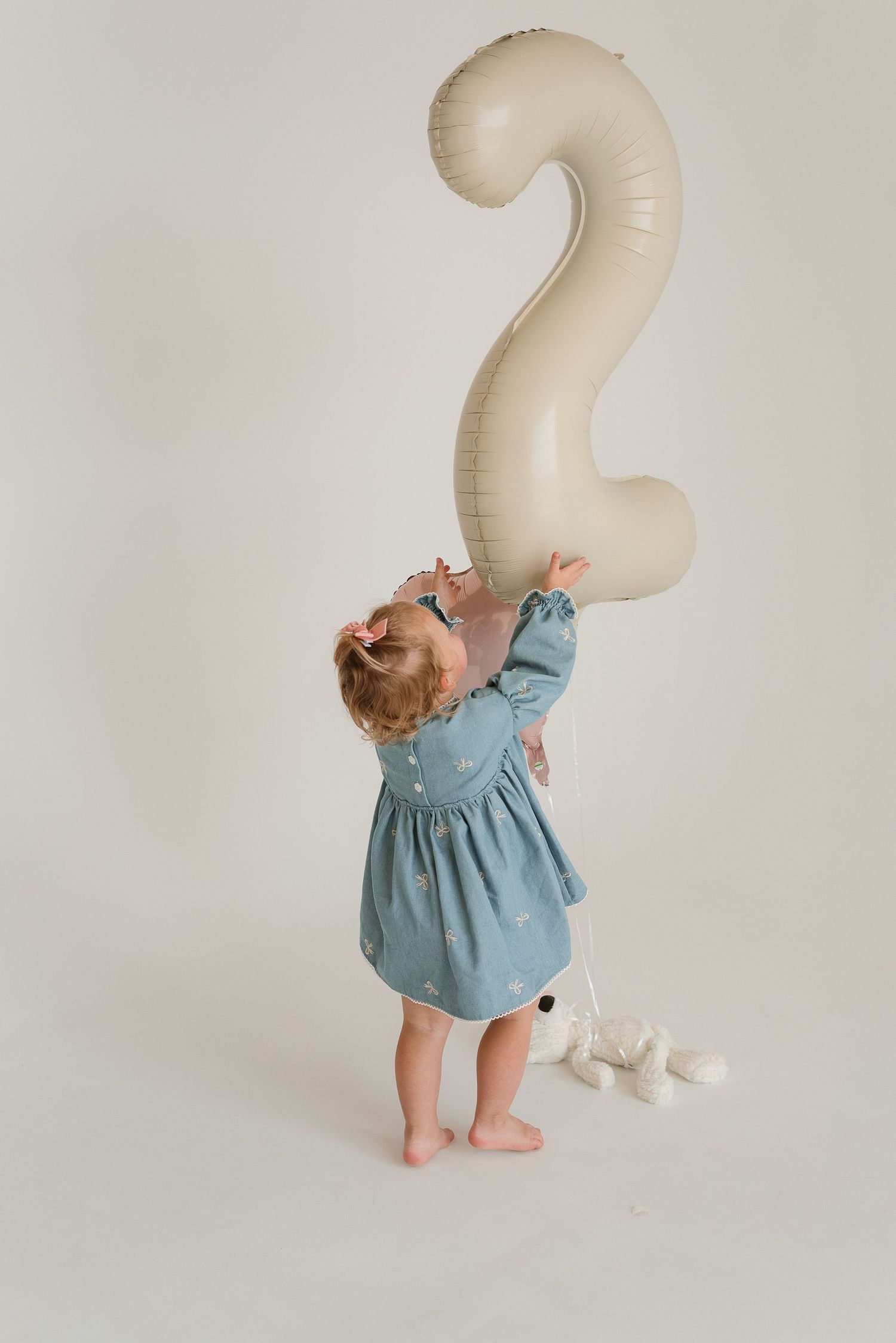 A toddler in dusty blue dress interacts with a large cream-colored number 2 balloon.
