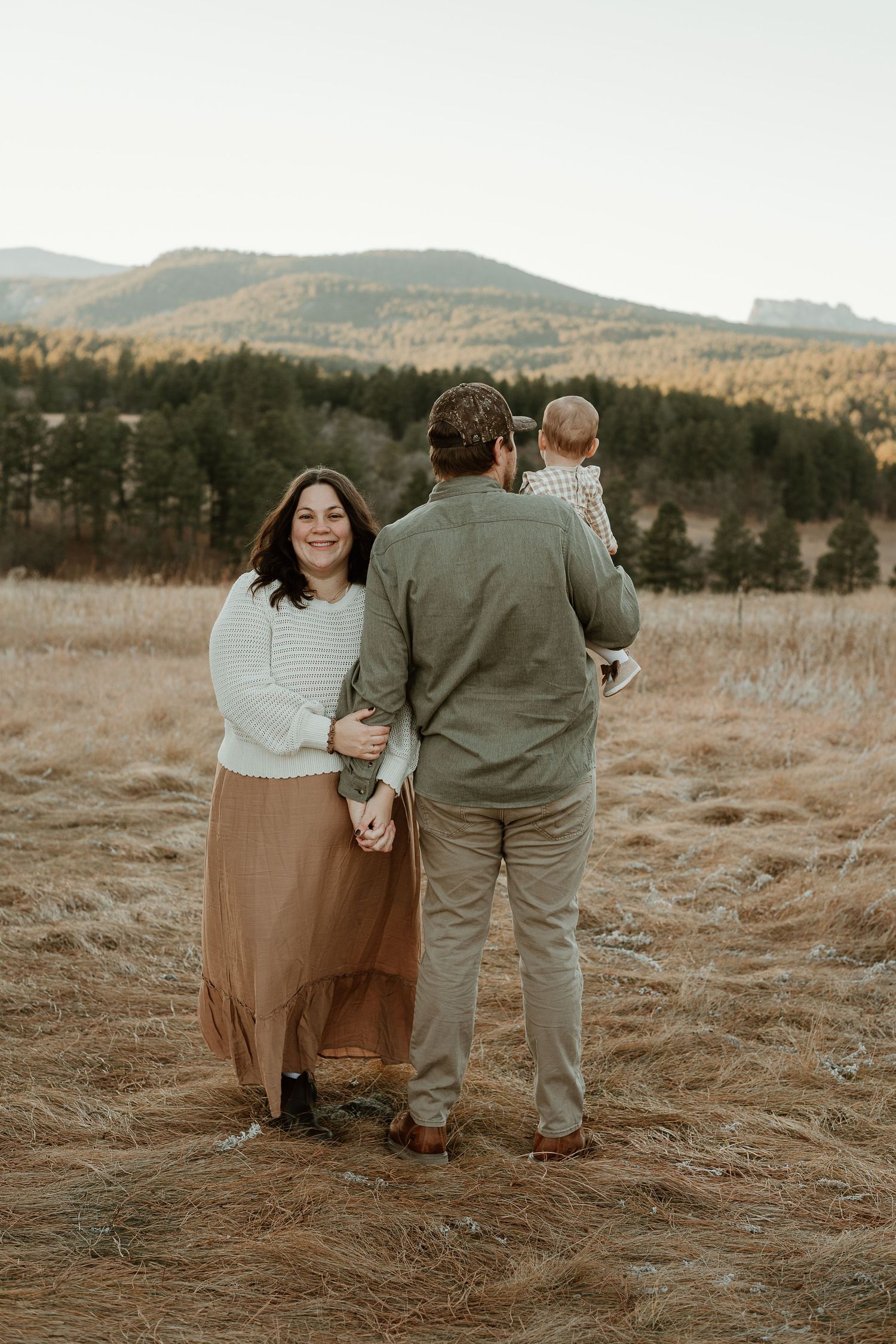 A couple stands together in a golden field at sunset with mountains and evergreen trees in the background.