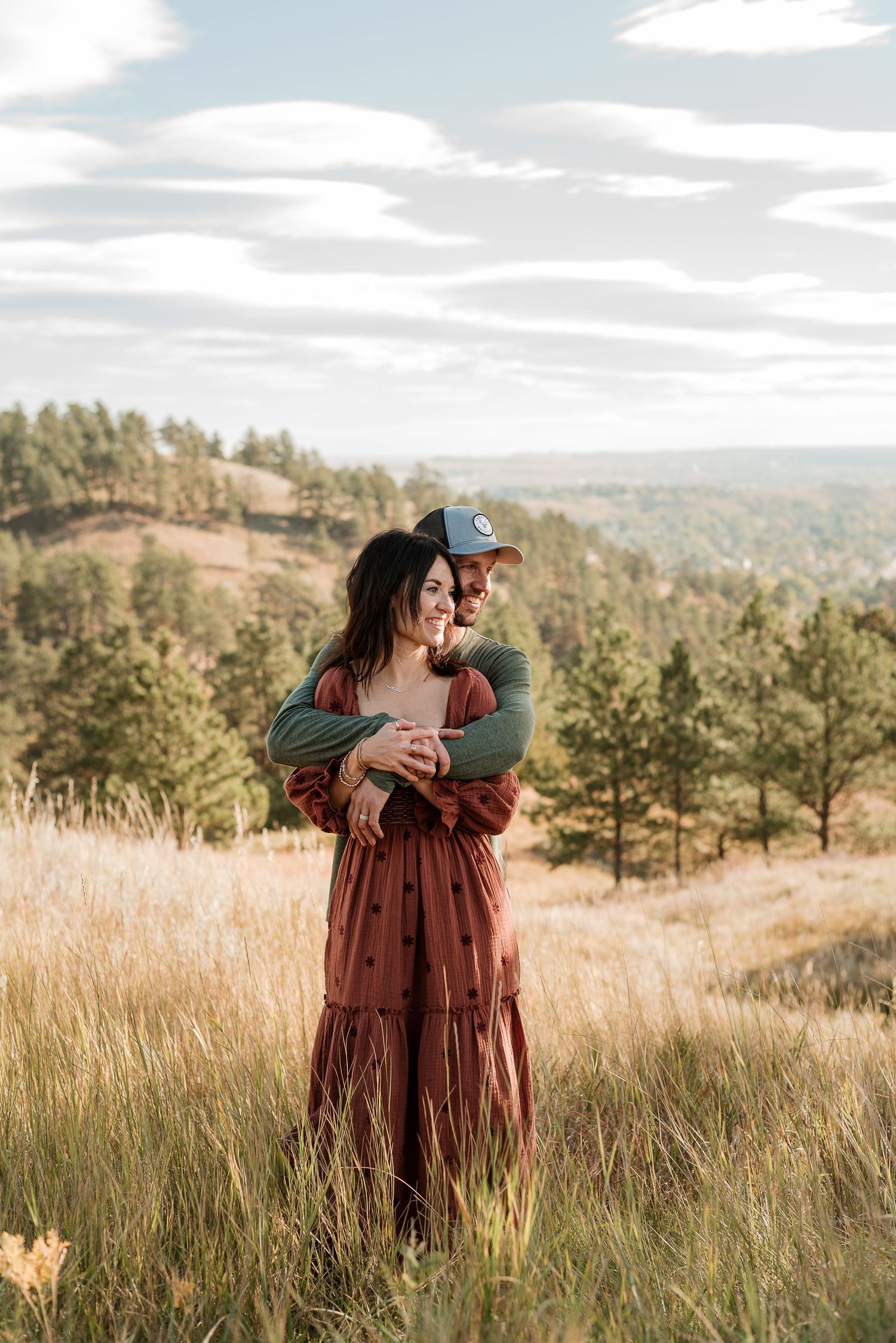 Couple embraces in tall grass field at sunset with rolling hills and pine trees in background.