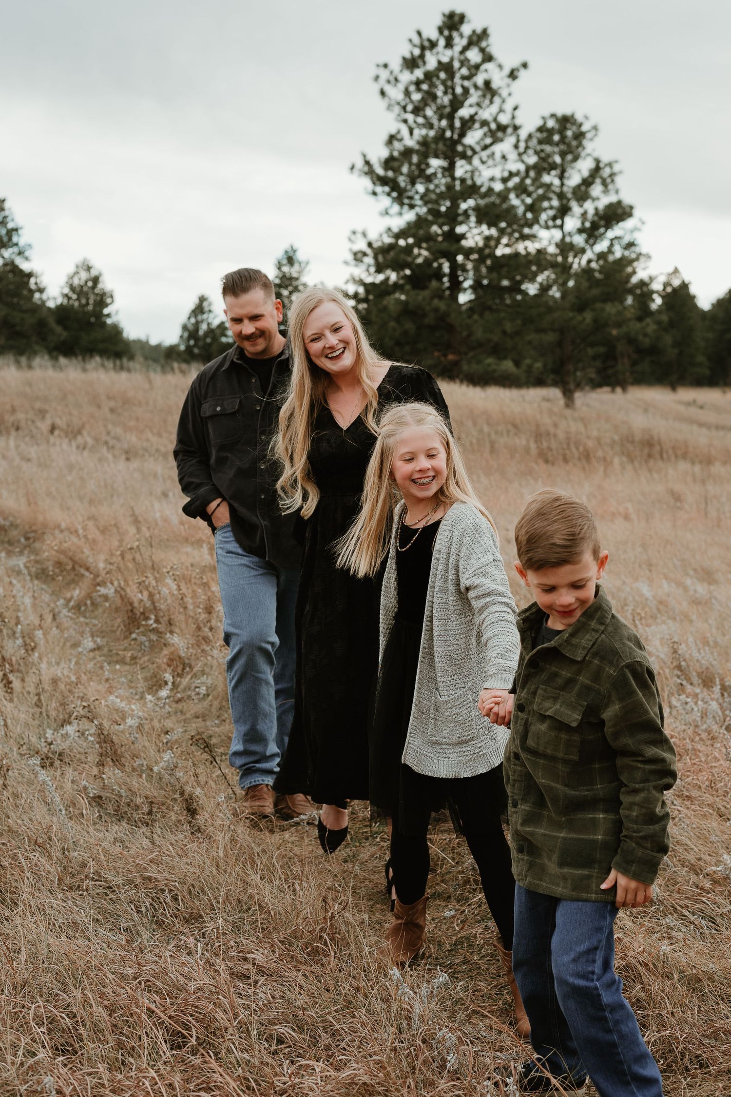 A family stands together in a line on a grassy hillside with evergreen trees in the background.